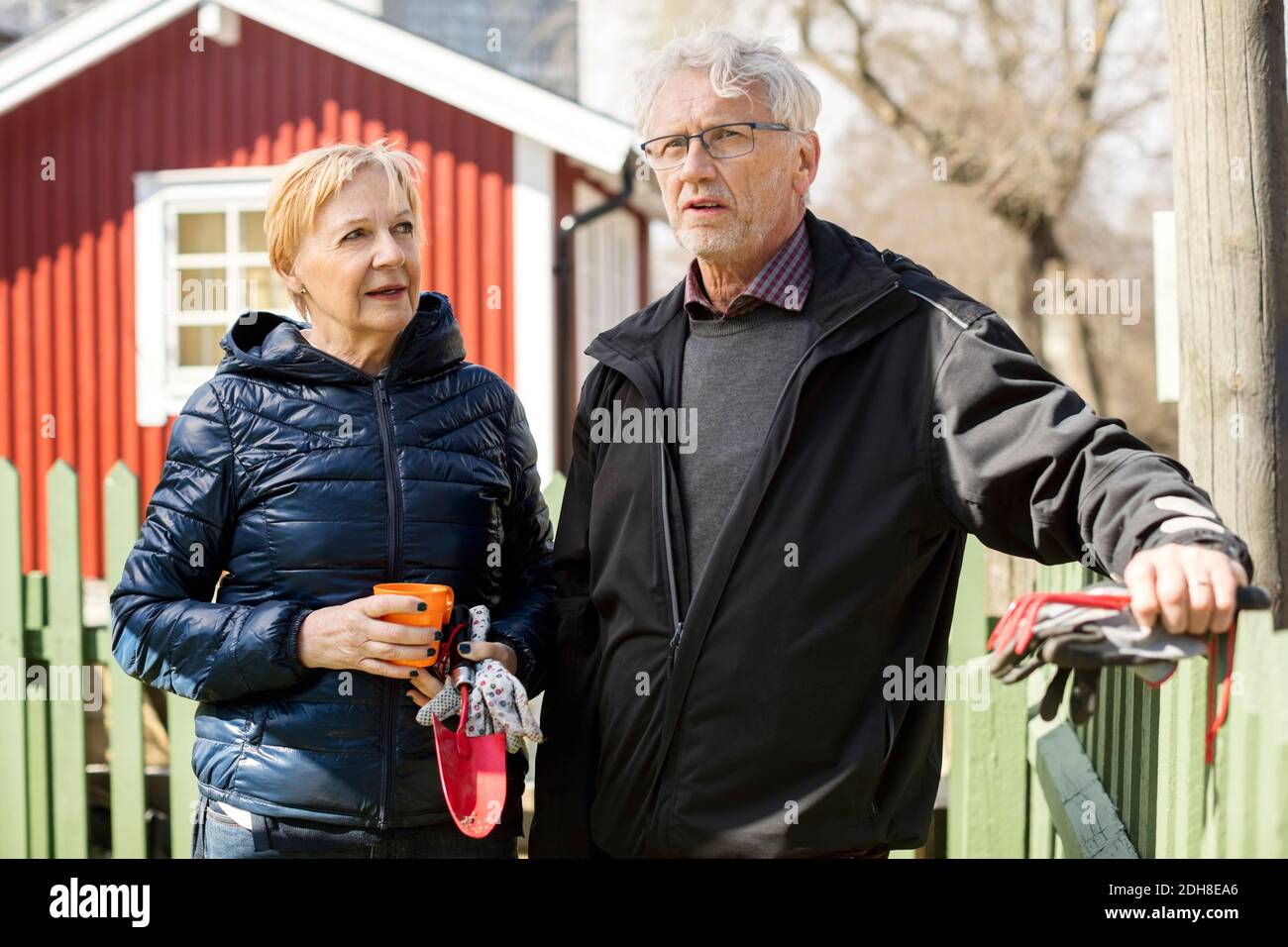 Lächelndes Seniorenpaar hält Gartengeräte, während es gegen den Zaun steht Im Winter Stockfoto