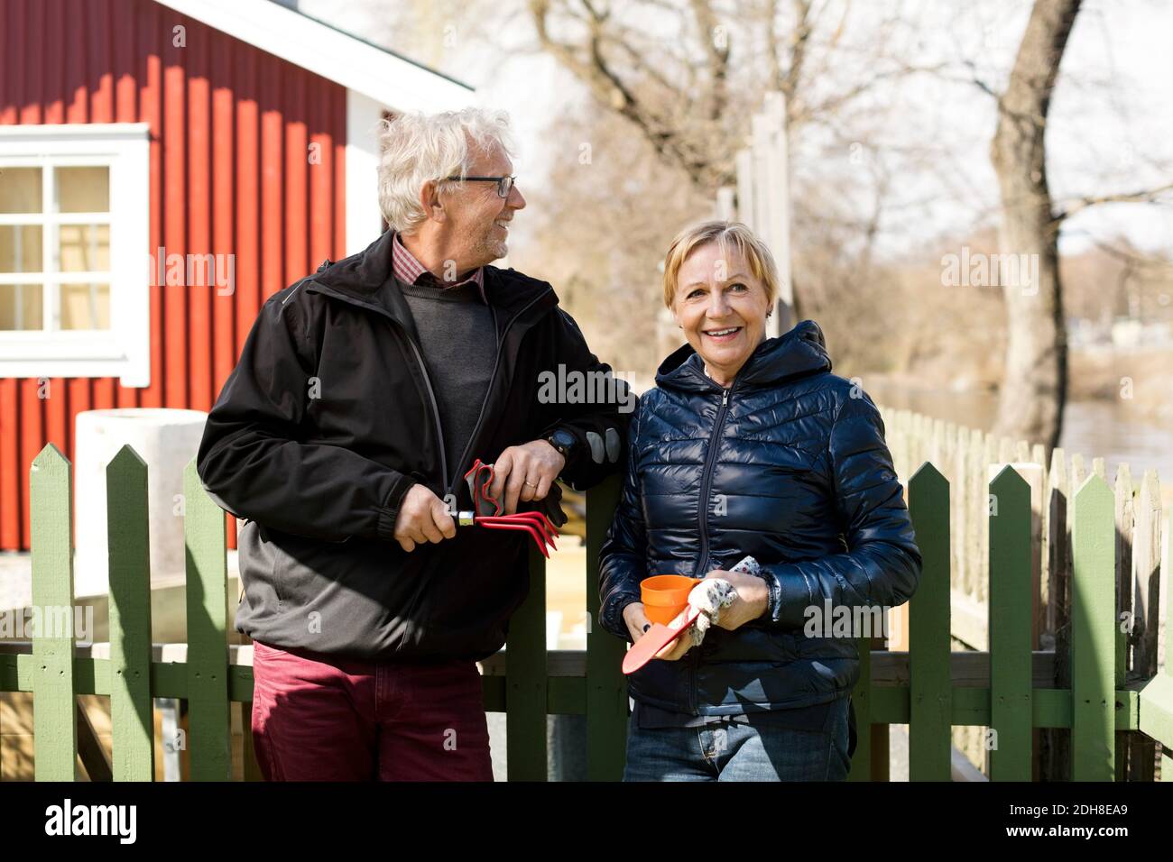 Lächelndes Seniorenpaar hält Gartengeräte, während es gegen den Zaun steht Im Winter Stockfoto