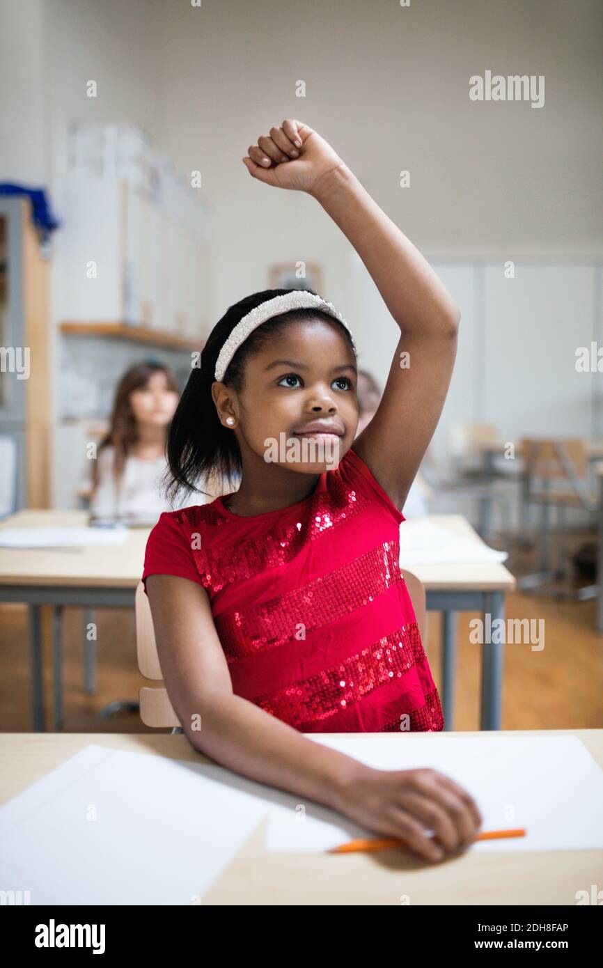 Konzentriertes Mädchen mit erhobener Hand am Schreibtisch im Klassenzimmer sitzen Stockfoto