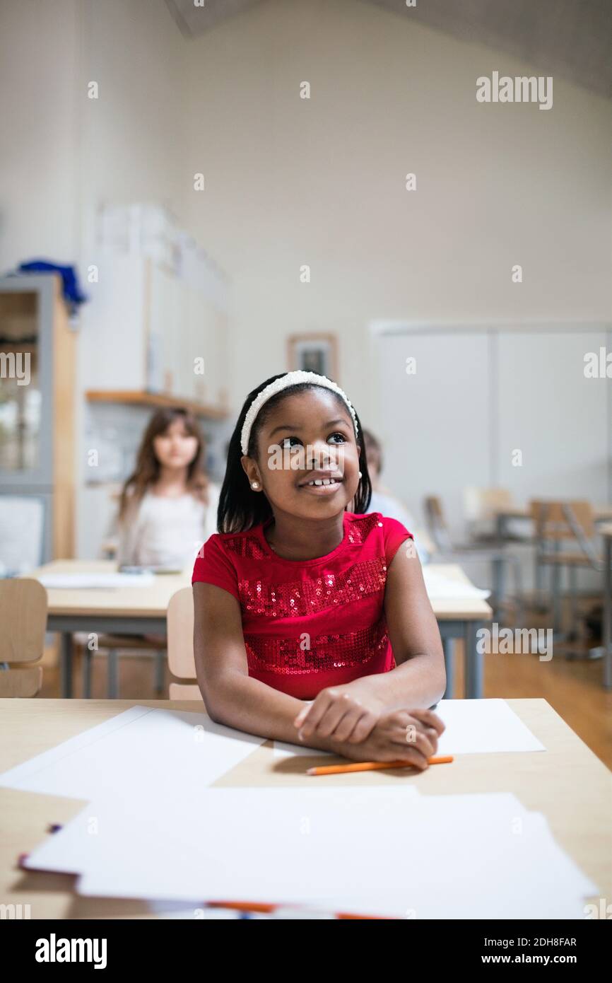 Lächelndes Mädchen hört zu, während es im Klassenzimmer am Schreibtisch sitzt Stockfoto