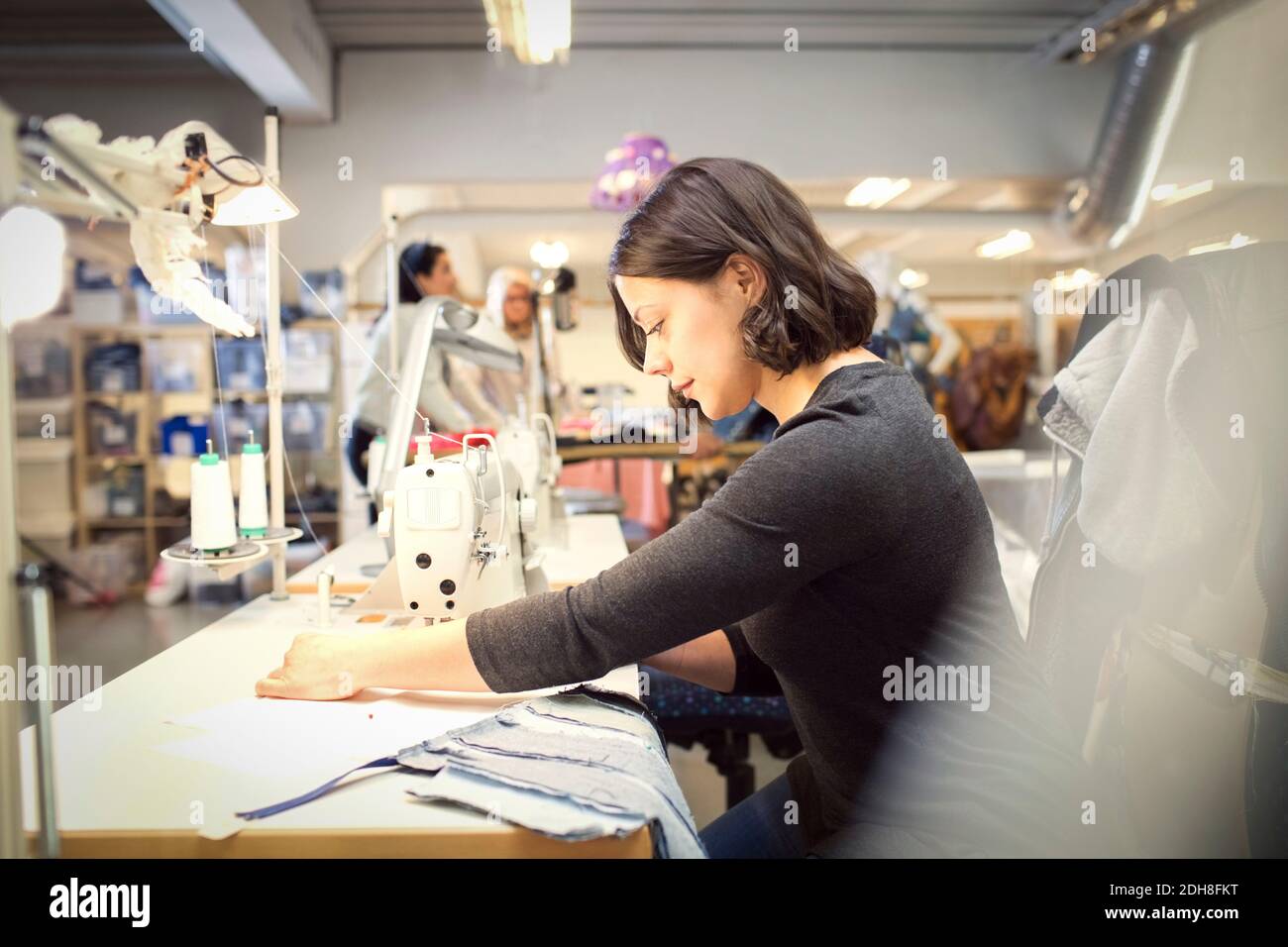 Seitenansicht der Frau nähen Stoff auf der Maschine, während Freiwillige Arbeiten in der Werkstatt Stockfoto