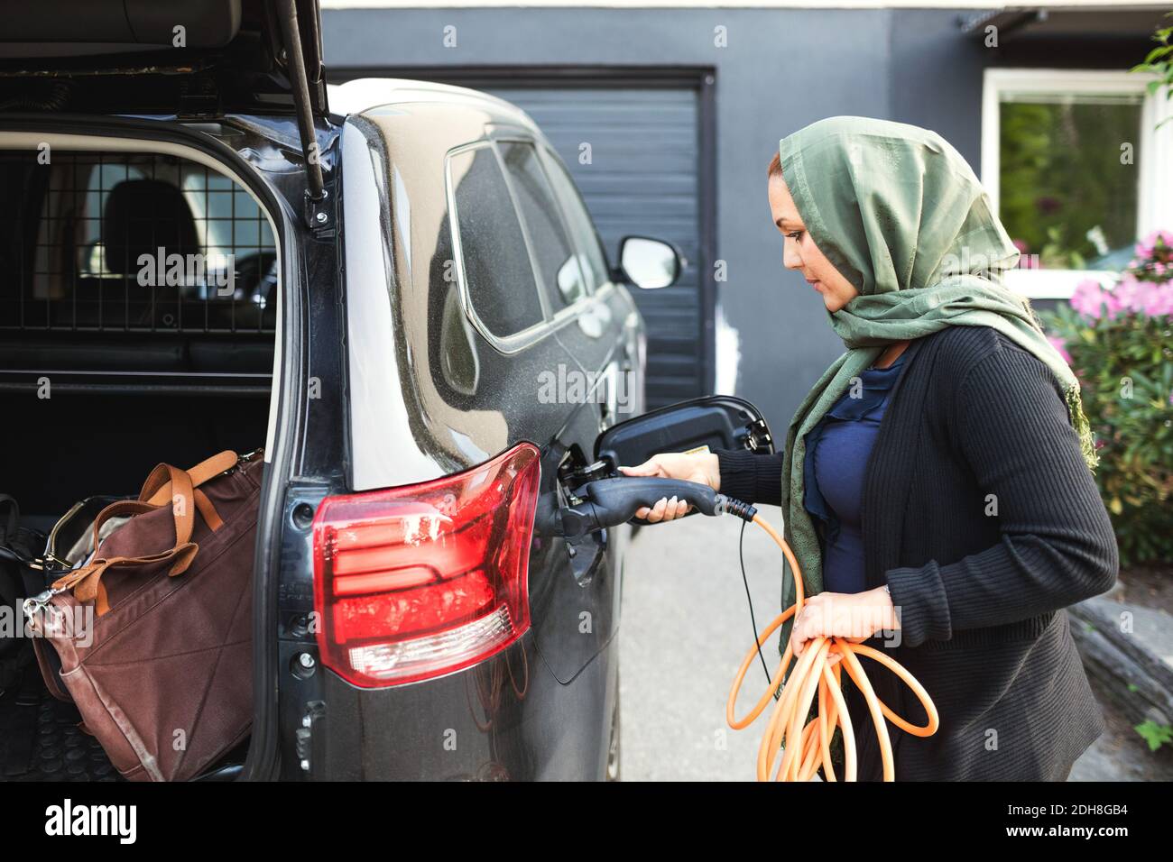 Seitenansicht einer Frau, die ein Elektroauto mit offenem Kofferraum lädt Während Sie vor dem Haus stehen Stockfoto