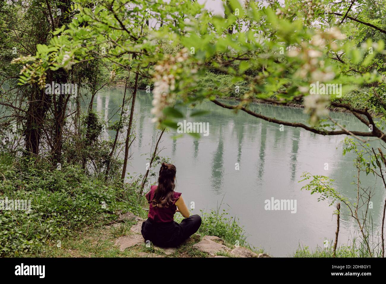 Frau, die in Lotusposition meditiert und am Ufer eines Flusses, dem Fluss Mincio, in Italien sitzt Stockfoto