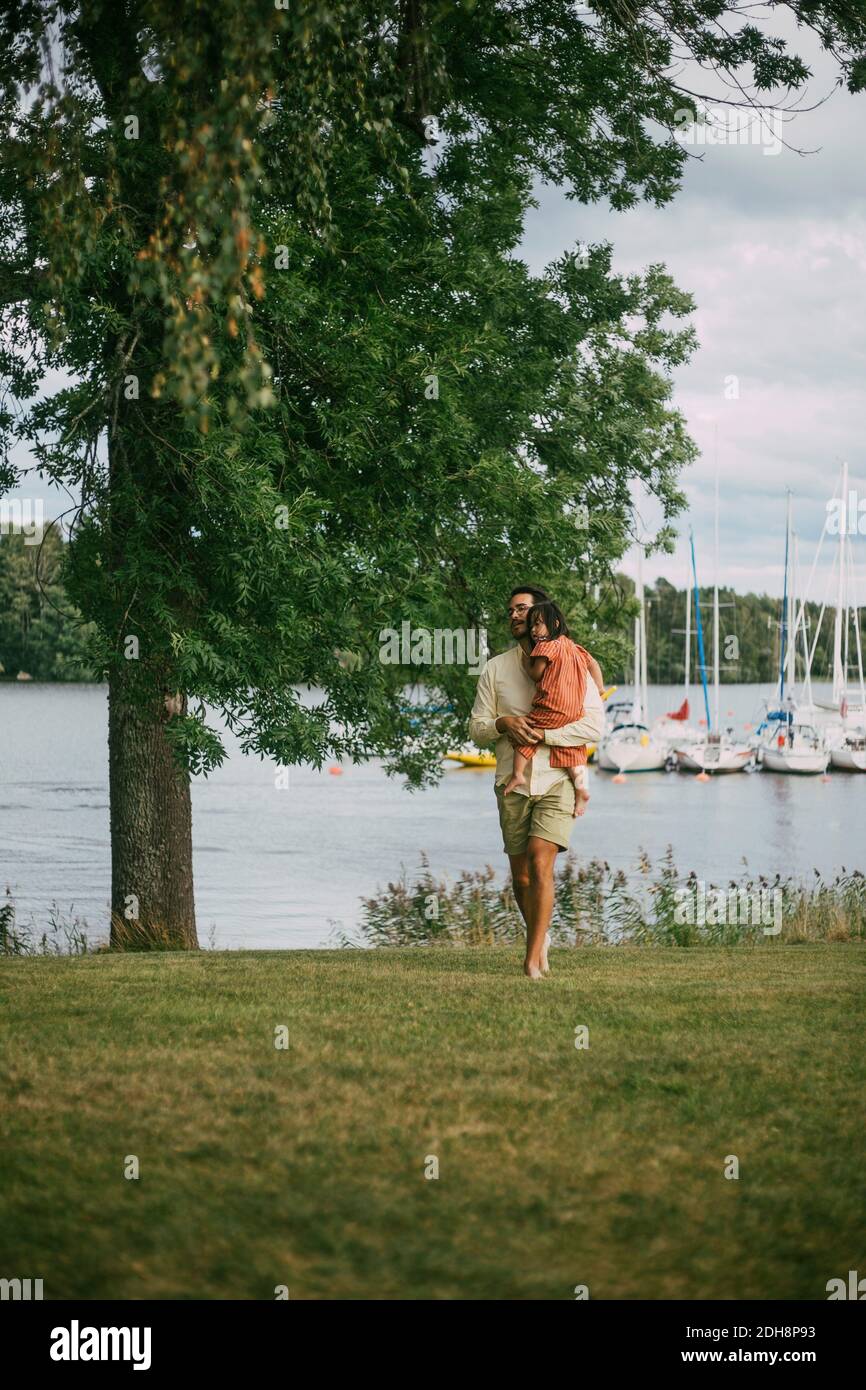 Vater trägt Tochter mit körperlicher Behinderung beim Gehen auf Gras Land gegen See im Sommer Stockfoto