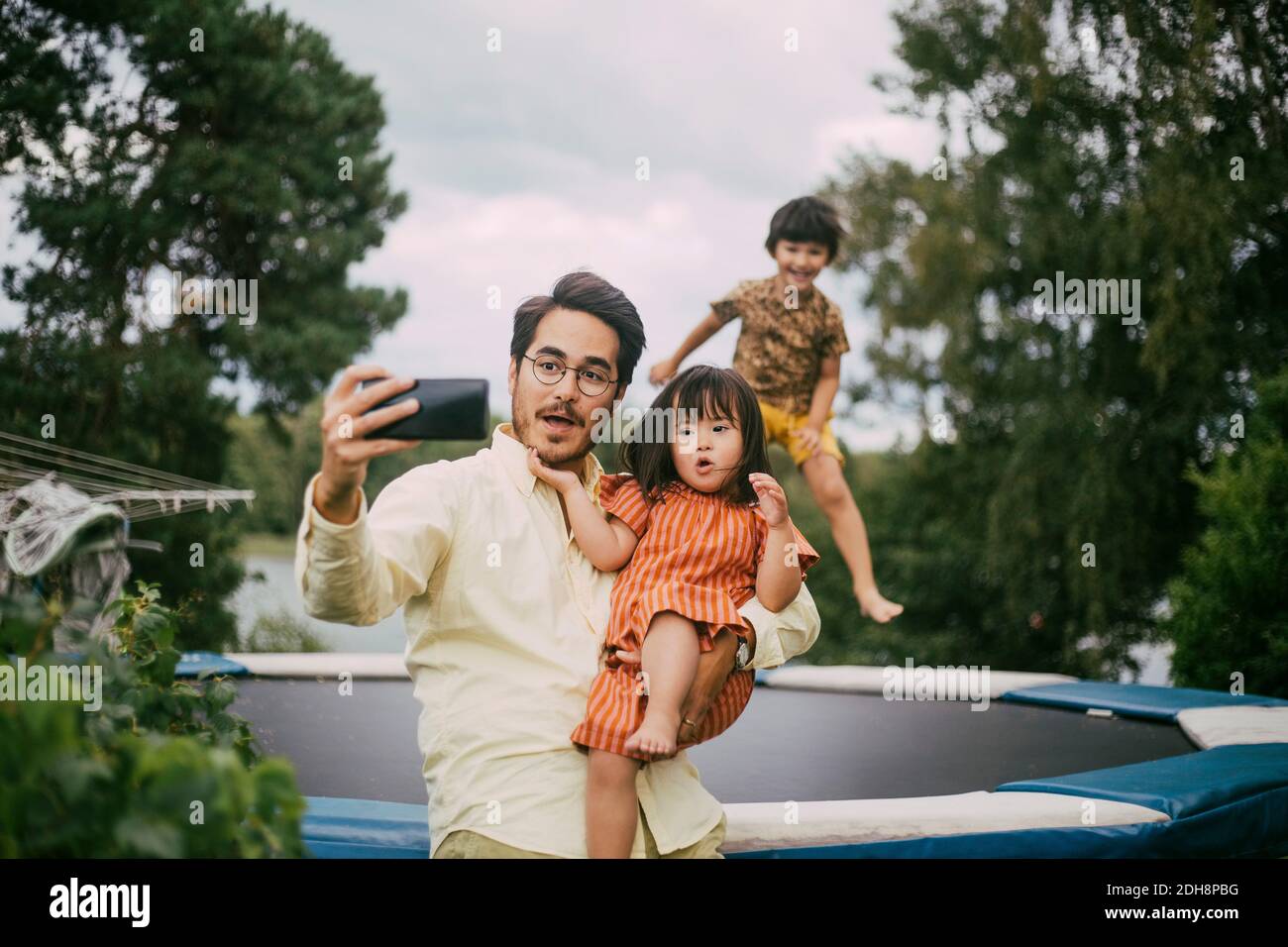 Lächelnder Vater trägt Syndrom Tochter während der Aufnahme Selfie gegen Mädchen springen auf Trampolin im Sommer Stockfoto