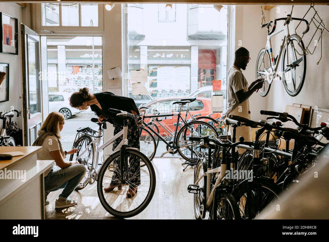 Weibliche Besitzerin Reparatur Fahrrad von älteren Kunden, während Kollegen arbeiten Auf dem Fahrrad in der Werkstatt Stockfoto