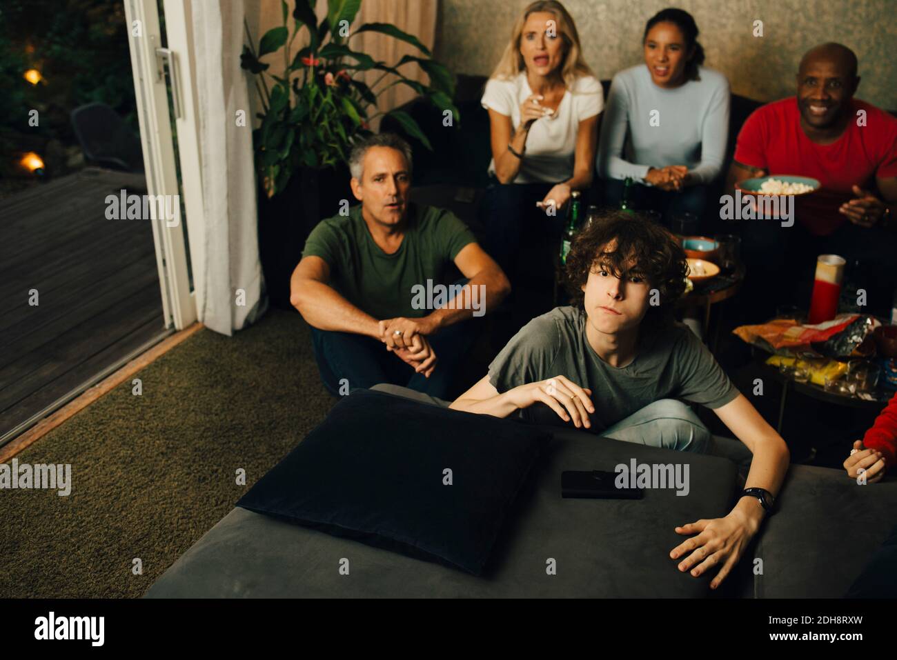 Männlicher Teenager auf dem Sofa beim Fernsehen während der Sportveranstaltung mit Familie bei Nacht Stockfoto