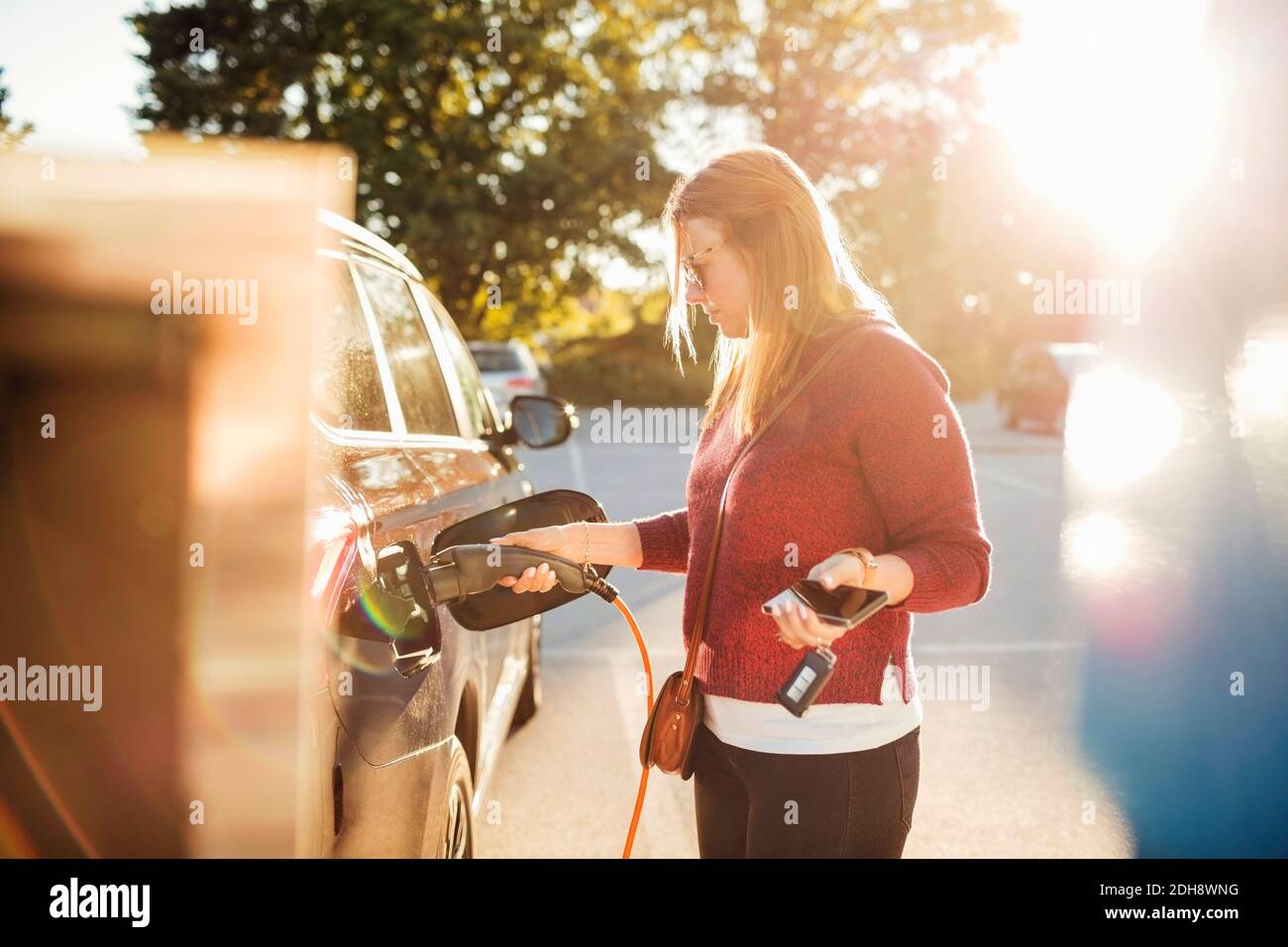 Frau hält Smartphone, während das Elektroauto an der Station geladen wird An sonnigen Tag Stockfoto