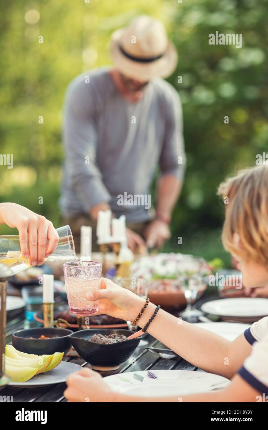 Kurze Hand des Teenagers Gießen Getränk in Glas gehalten Von Bruder über Esstisch im Hinterhof Stockfoto