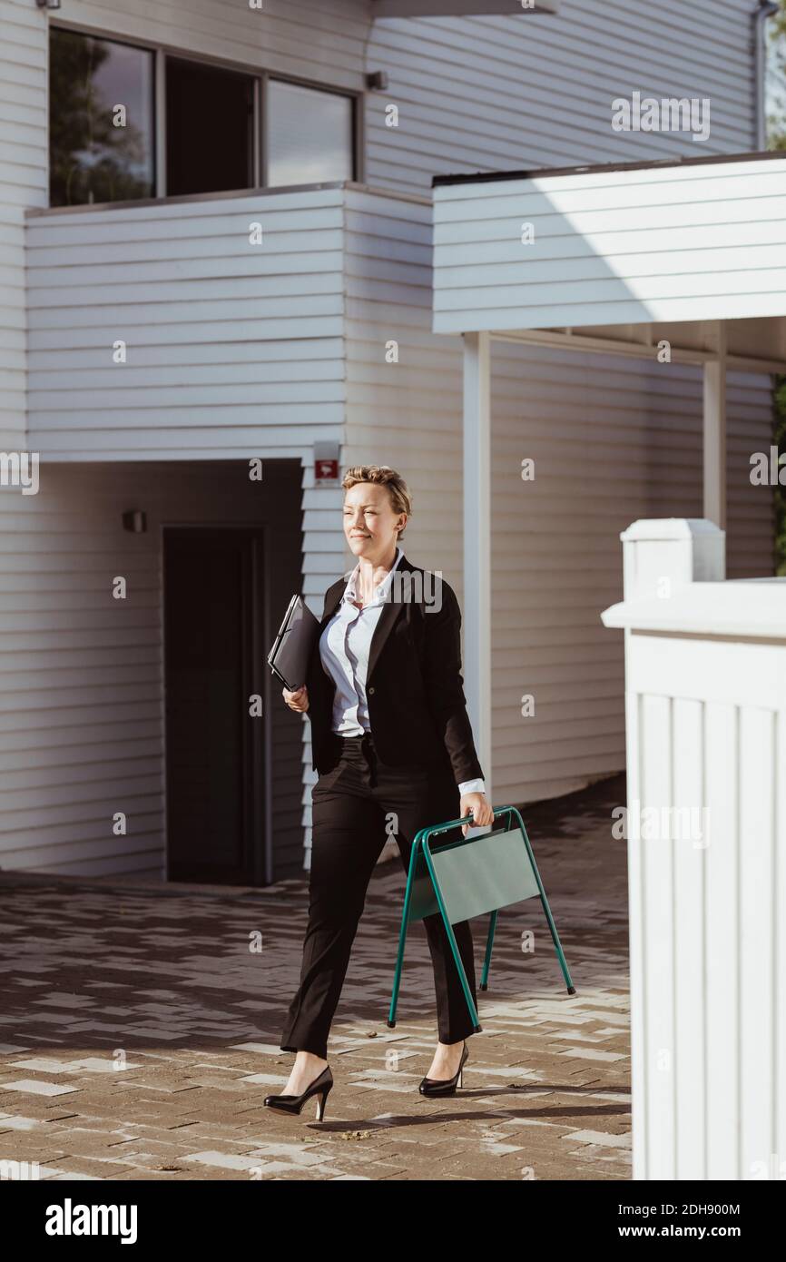 Lächelnd weibliche Immobilienmakler wegschauen, während zu Fuß gegen Haus Stockfoto