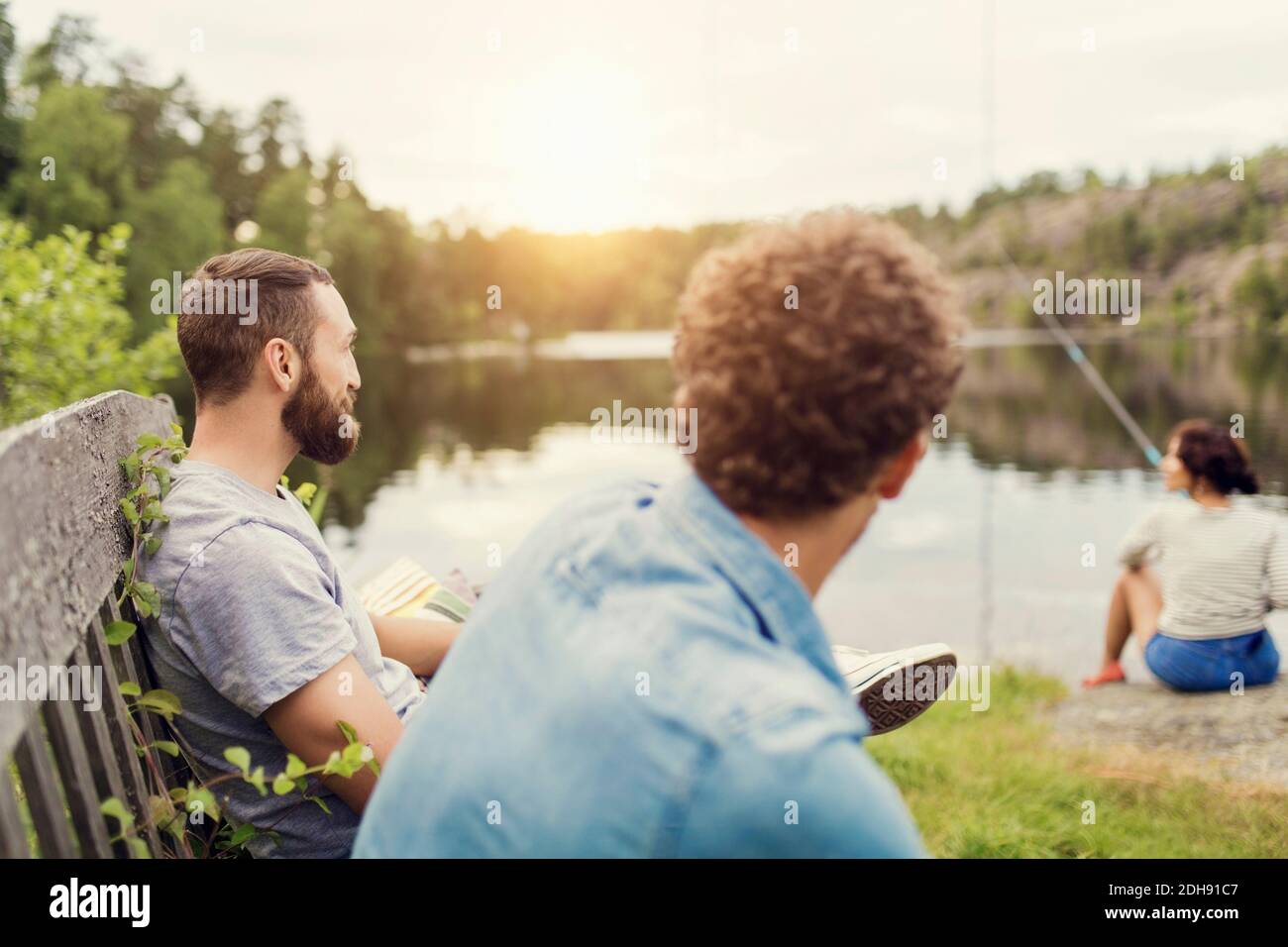 Männer sitzen auf Holzbank, während weibliche Freundin beim Angeln Seeufer Stockfoto