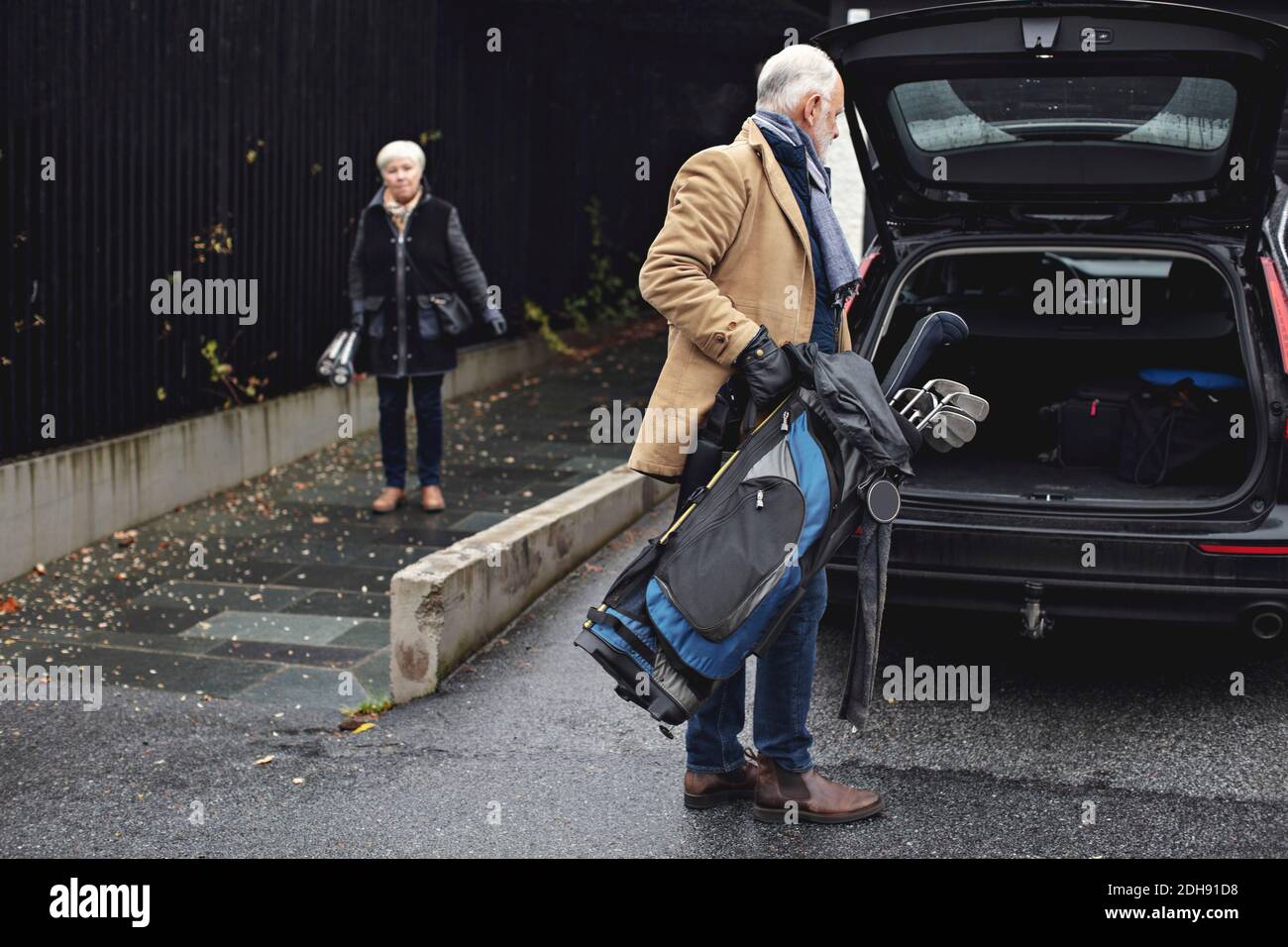 Älterer Mann hält Golftasche am Kofferraum des Autos, während sie weiblich ist Partner steht im Winter auf dem Bürgersteig Stockfoto