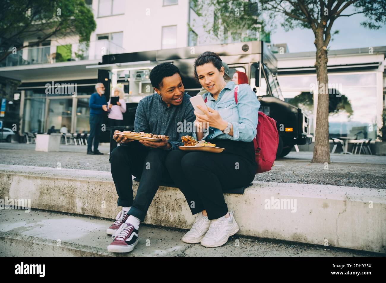 Lächelnde Freunde teilen Smartphone, während sie mit Essen gegen sitzen Food Truck in der Stadt Stockfoto
