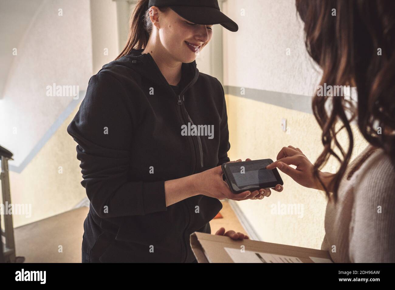 Lächelnde Lieferung Frau hält Telefon, während der Kunde während der Unterzeichnung zu Hause Lieferung Stockfoto