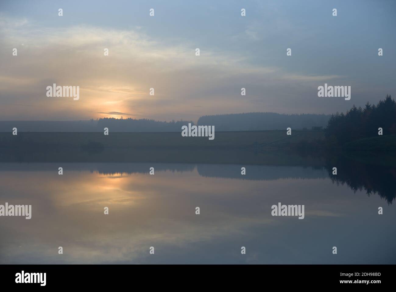 Dammwand wirkt als Symmetrielinie in dieser reflektierten Landschaft. Dunstig Sonnenuntergang über dem ruhigen Wasser der Redmires Reservoirs. Kalter Winter in der Abenddämmerung. Stockfoto