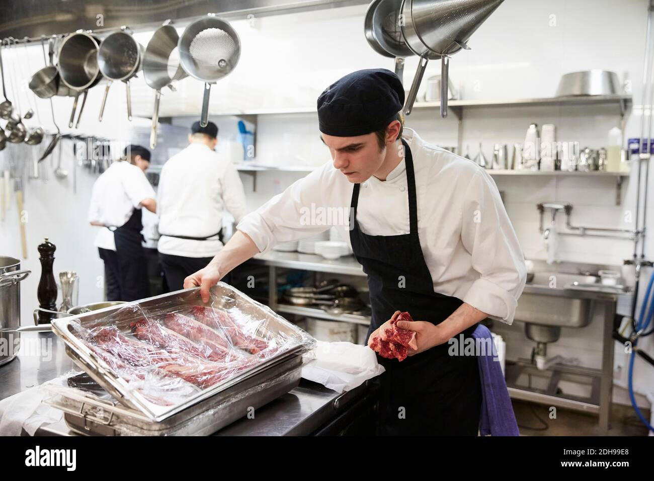 Männlicher Koch untersucht Tablett mit Fleisch mit Kunststoff in verpackt Gewerbliche Küche Stockfoto