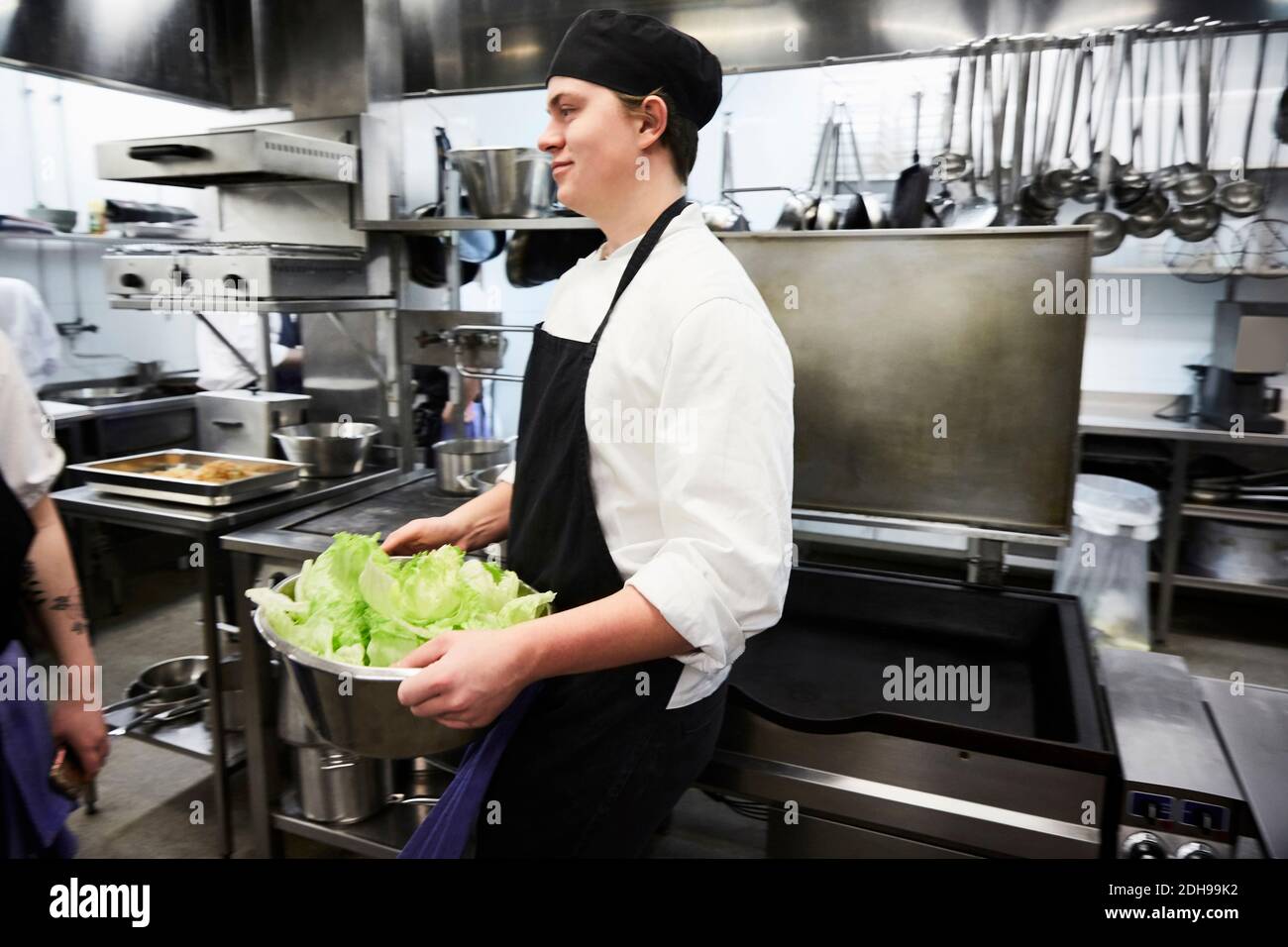 Seitenansicht des männlichen Chefstudenten, der Behälter mit Gemüse hält In der kommerziellen Küche Stockfoto