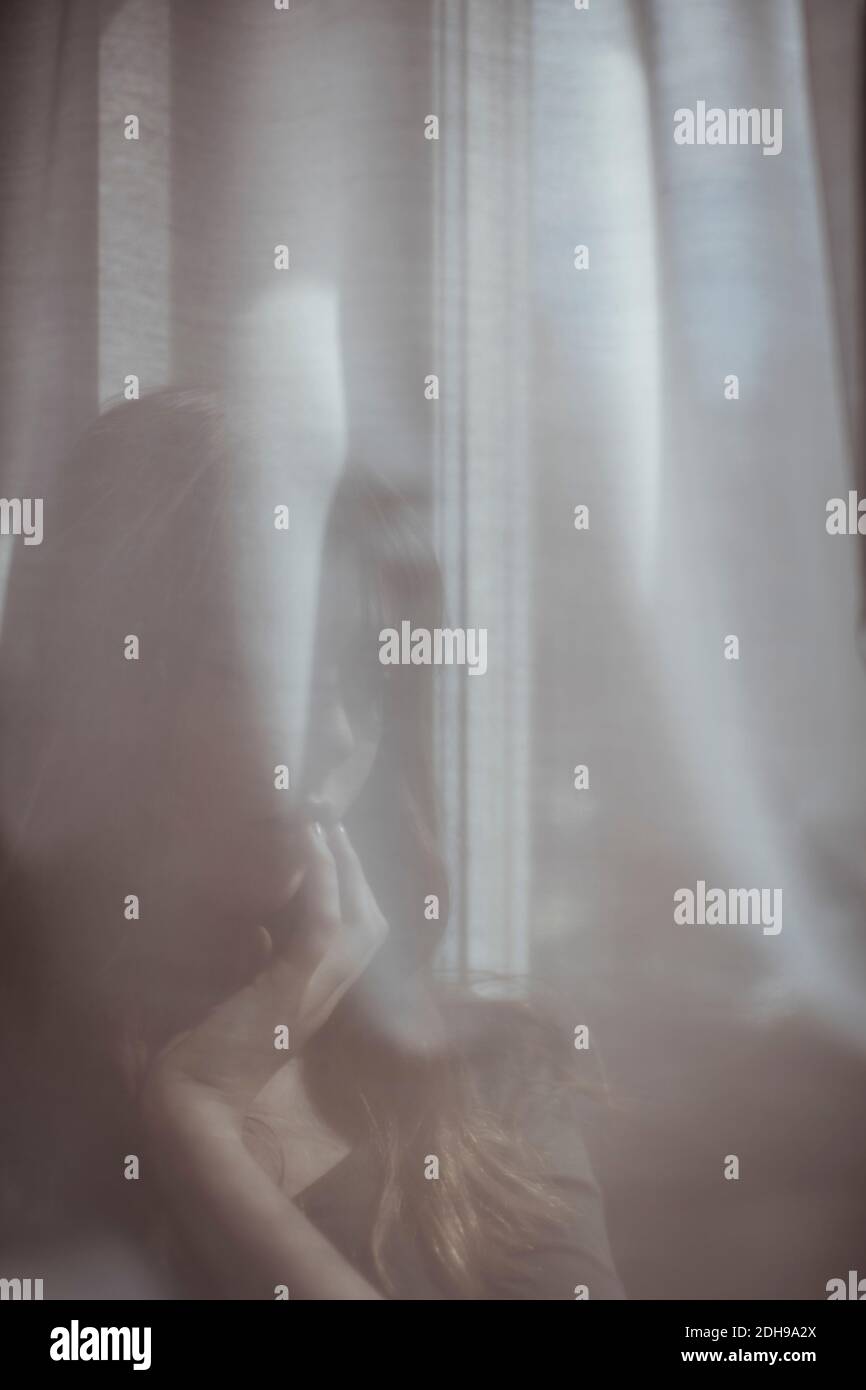 Frau versteckt sich hinter weißem Fenstervorhang Stockfoto