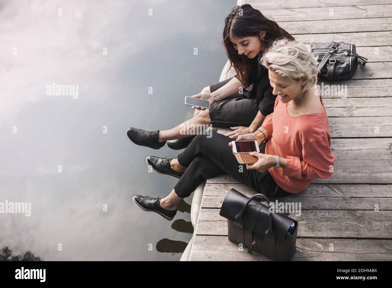 High-Angle-Ansicht von glücklichen Geschäftsfrauen mit Smartphone, während Sitzen am Pier am See Stockfoto