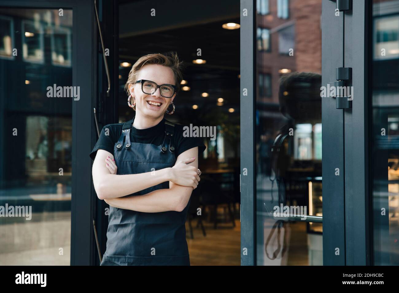 Portrait eines lächelnden androgynen Besitzers, der mit gekreuzten Armen steht Eingang des Cafés Stockfoto