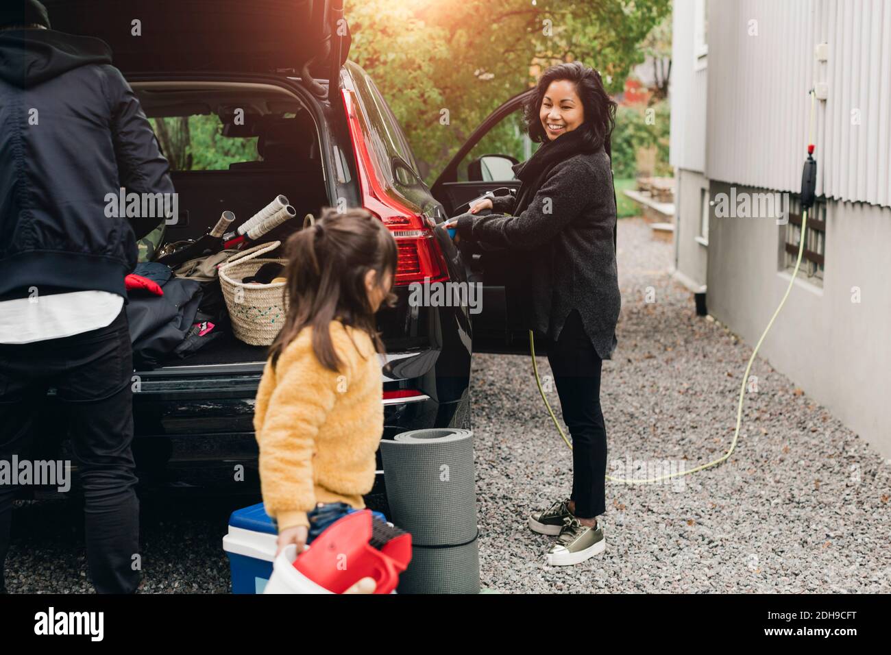 Lächelnde Frau lädt das Auto, während die Familie Gepäck im Kofferraum lädt Stockfoto
