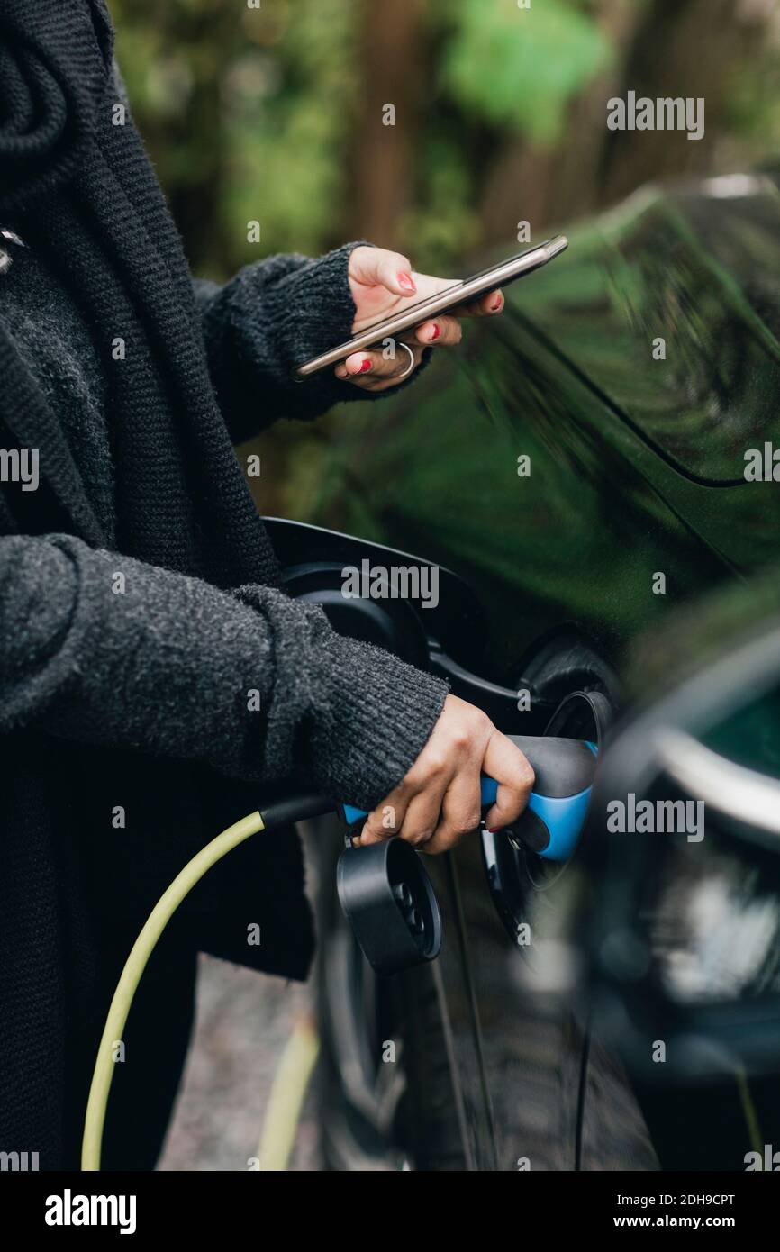 Mittelteil der mittleren erwachsenen Frau, die während des Ladevorgangs ein Mobiltelefon verwendet Elektroauto Stockfoto