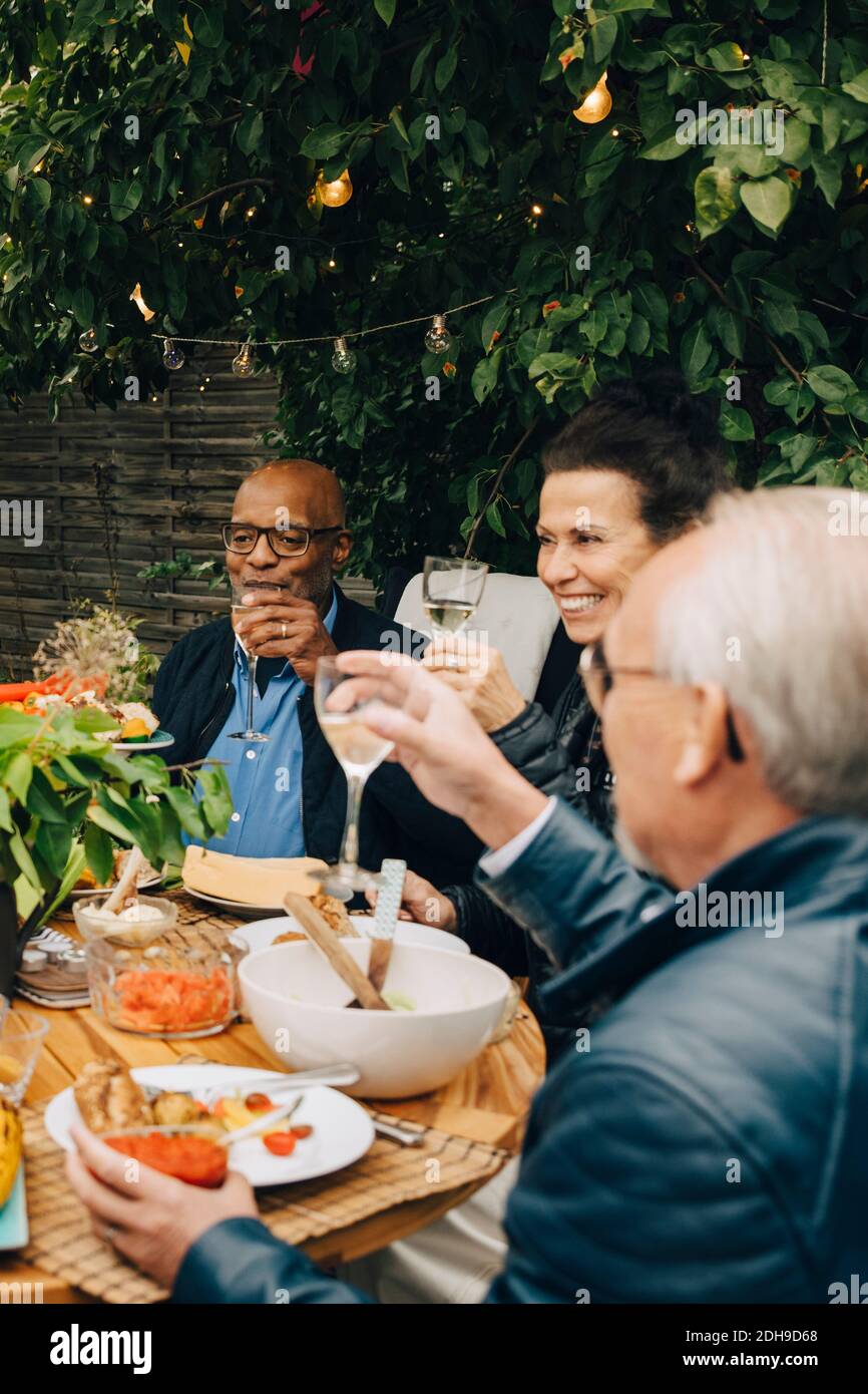Lächelnde ältere Frau, die mit männlichen Freunden sitzt, während sie Toast aufwirft Am Esstisch während der Dinner-Party im Hinterhof Stockfoto