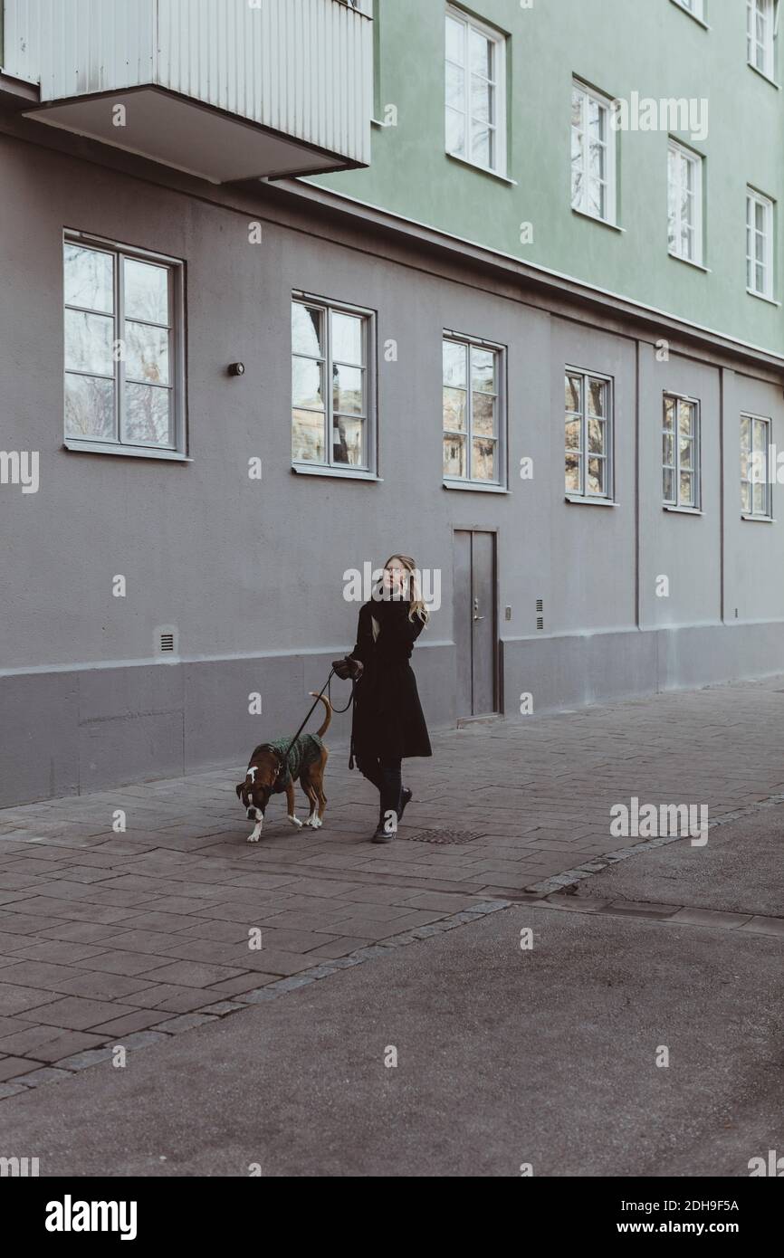Die ganze Länge der jungen Frau, die mit Hund auf dem Fußweg läuft Durch Gebäude in der Stadt Stockfoto