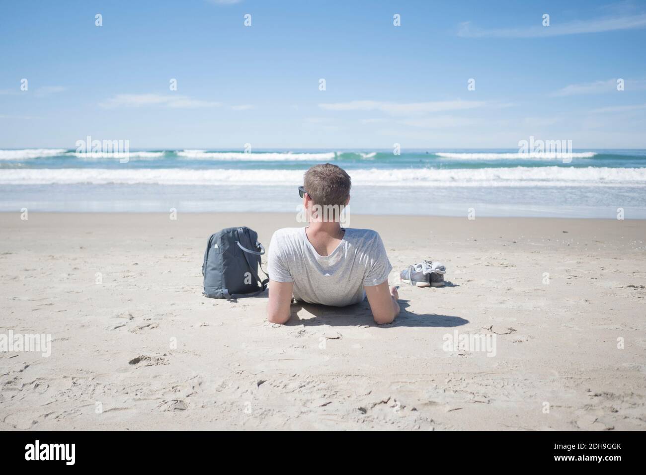 Rückansicht des Mannes, der mit dem Rucksack am Strand relaxt Sonniger Tag Stockfoto
