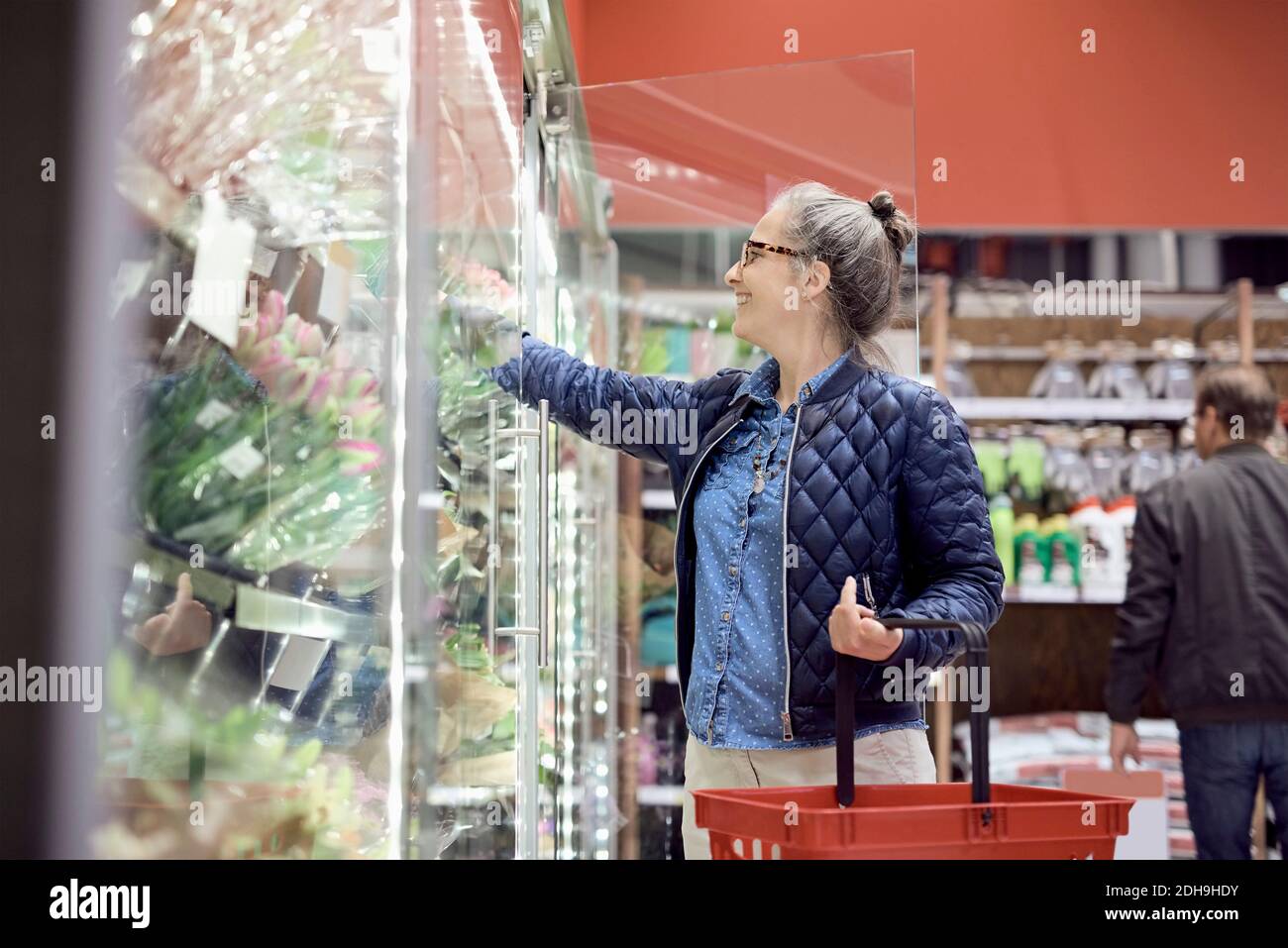 Lächelnde Frau, die Blumen kauft, während sie den Einkaufskorb im Supermarkt hält Stockfoto