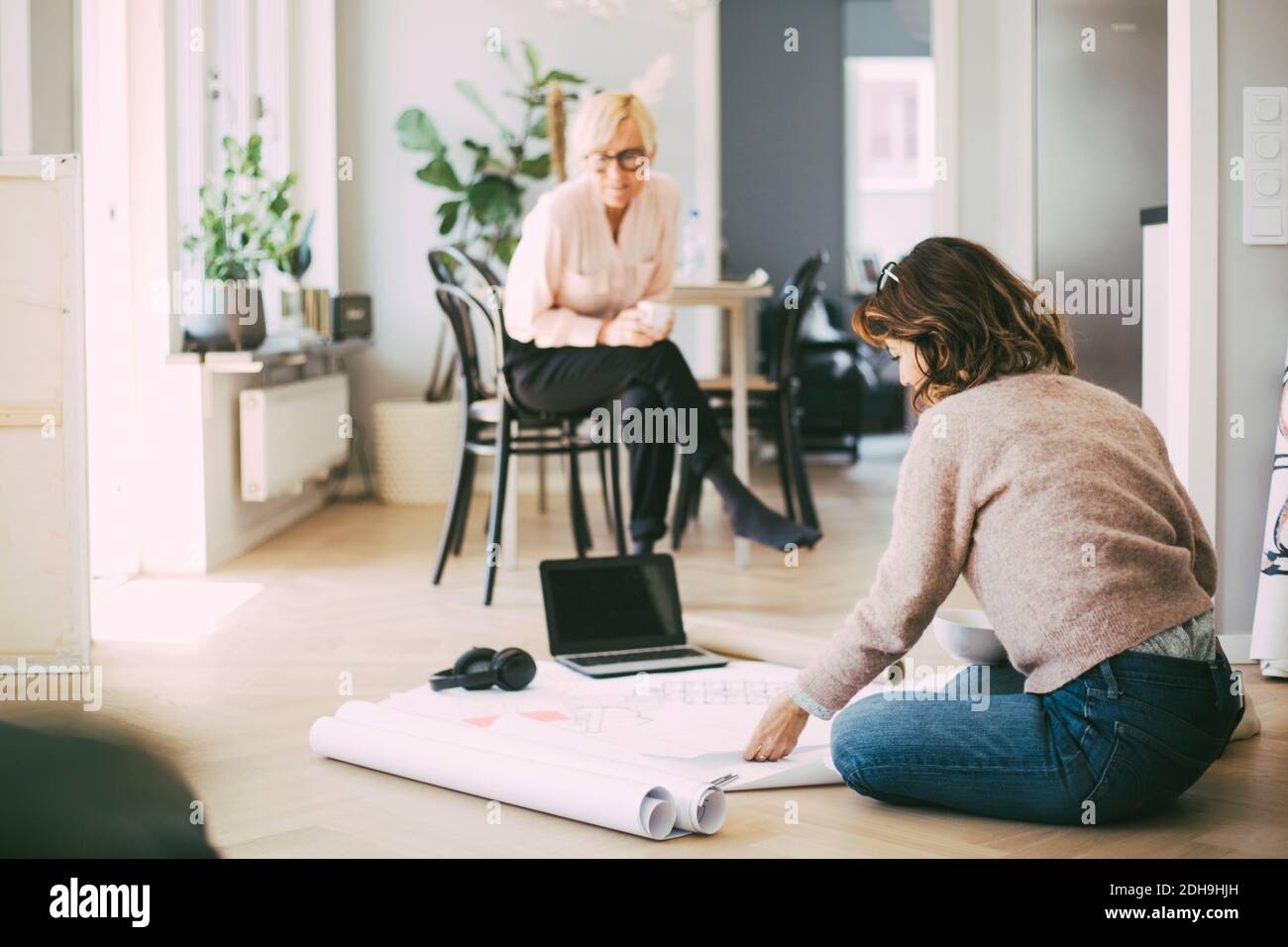 Frau, die auf dem Boden sitzt und mit einer Kollegin über die Blaupause diskutiert Zu Hause Stockfoto