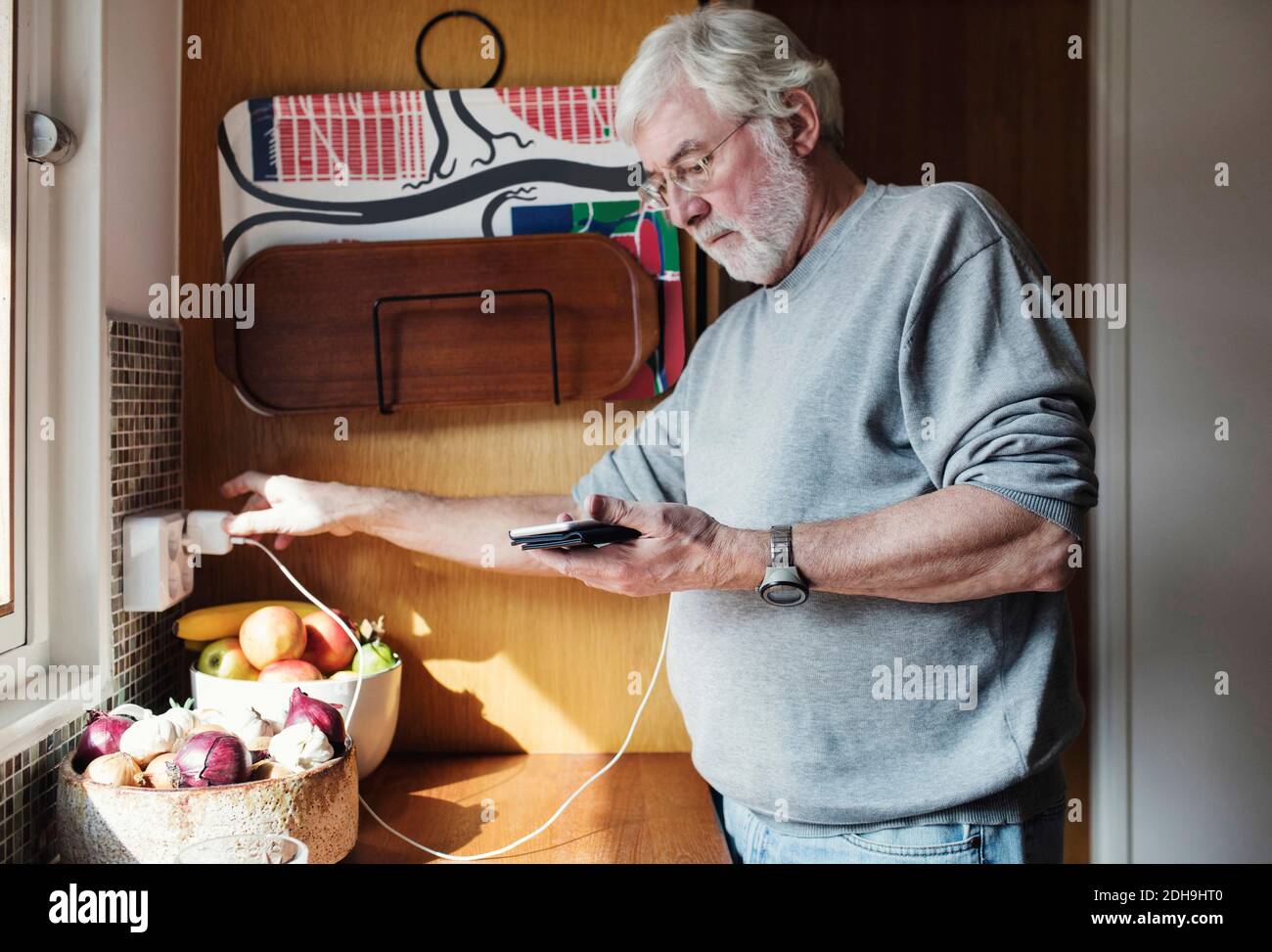 Senior Mann hält Handy, während das Ladegerät an die Stromversorgung angeschlossen wird Steckdose in der heimischen Küche Stockfoto
