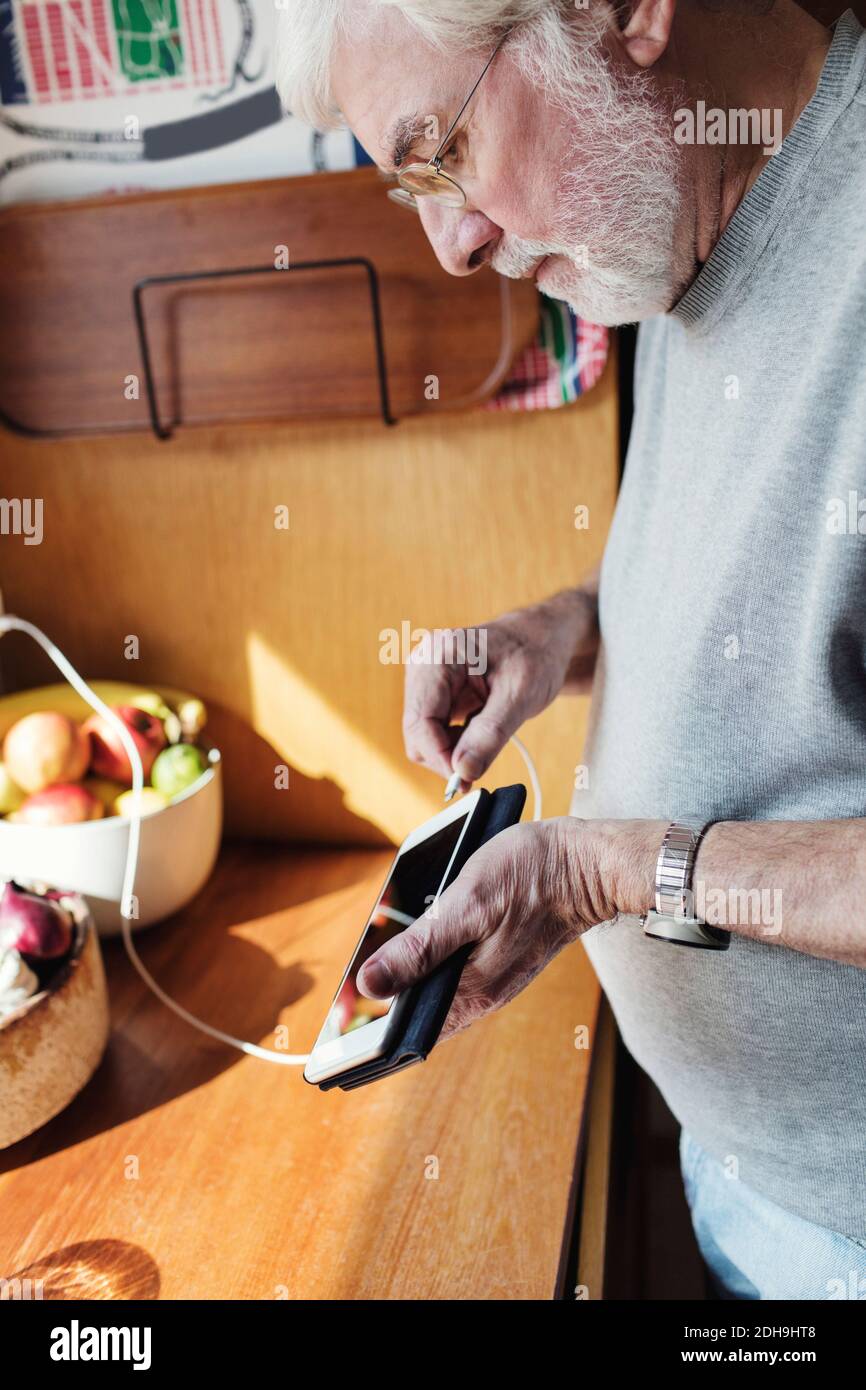 Älterer Mann, der das Kabel an das Mobiltelefon an der Küchentisch anschlusstelefon An sonnigen Tag Stockfoto