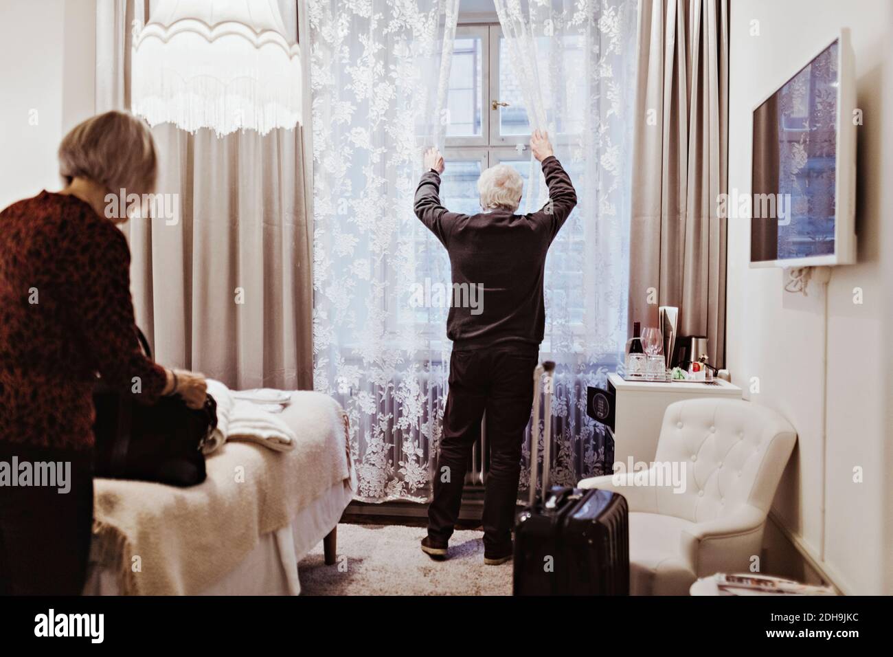 Älterer Mann, der die Vorhänge öffnete, während eine Frau die Tasche im Hotel hielt Zimmer Stockfoto