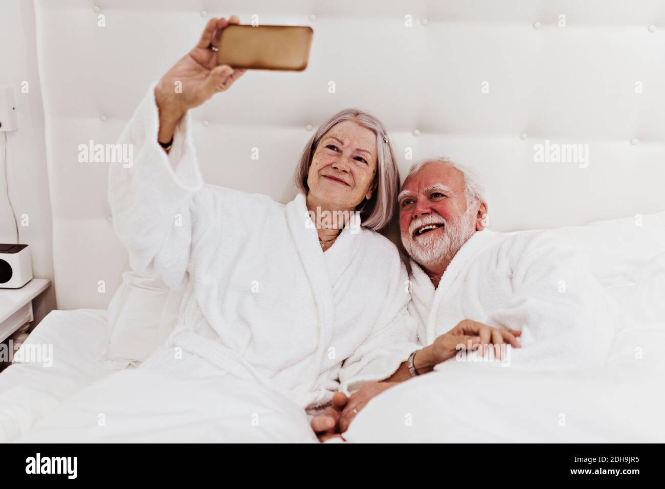 Lächelnde ältere Frau, die mit dem Mann Selfie über das Mobiltelefon nimmt Im Hotelzimmer auf dem Bett liegen Stockfoto