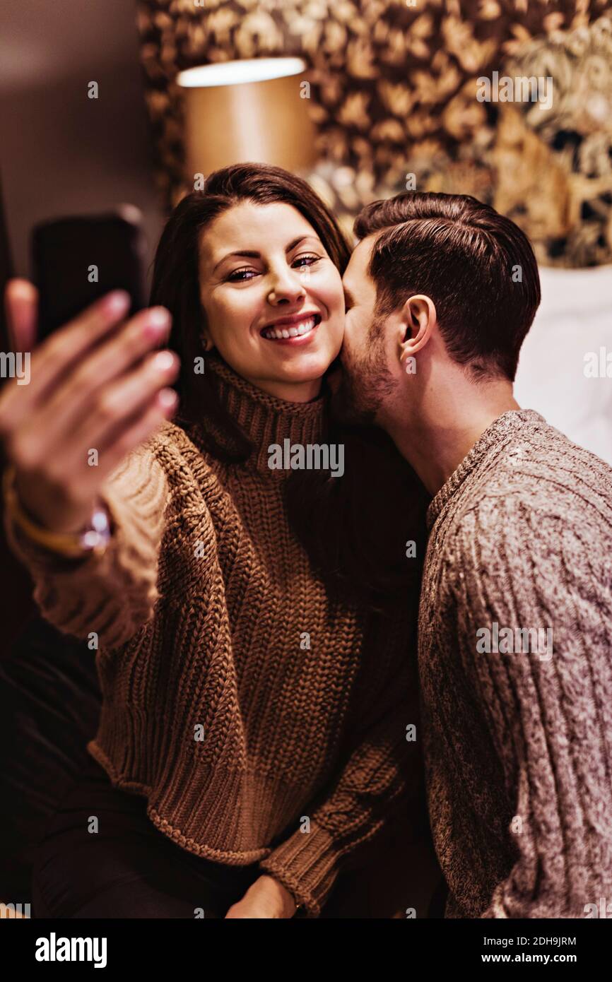 Mann küsst, während lächelnde Freundin Selfie im Hotelzimmer nimmt Stockfoto