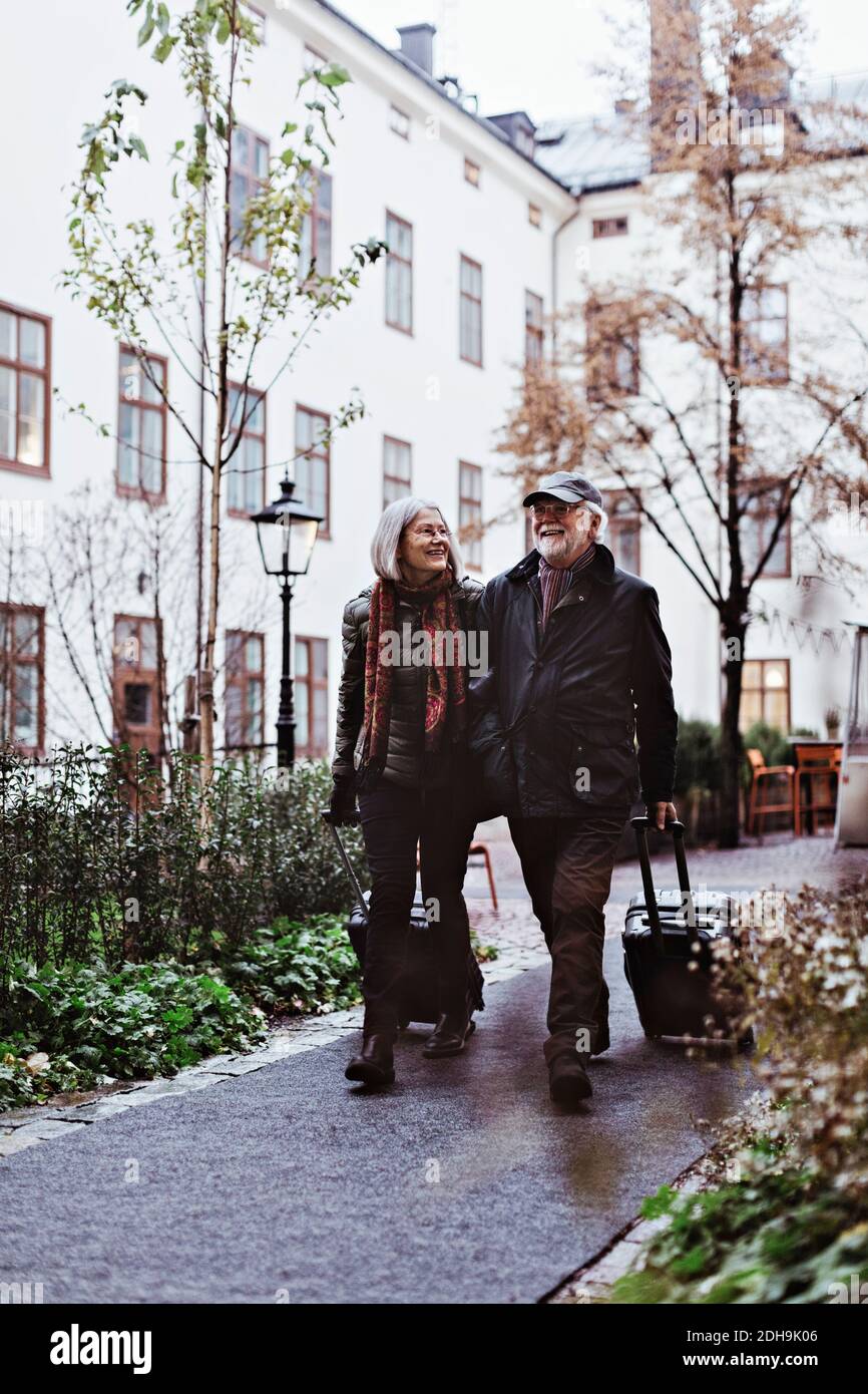 Ein glückliches Ehepaar, das mit dem Gepäck vor dem Hotel auf dem Fußweg läuft Stockfoto