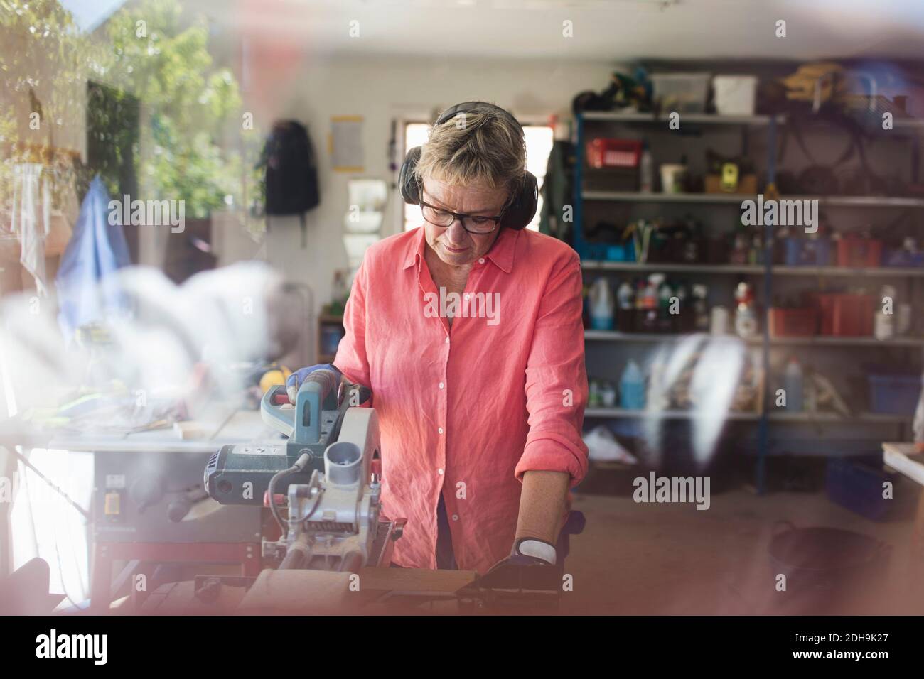 Ältere Frau mit Kreissäge in der Werkstatt durch Glas gesehen Angezeigt Stockfoto