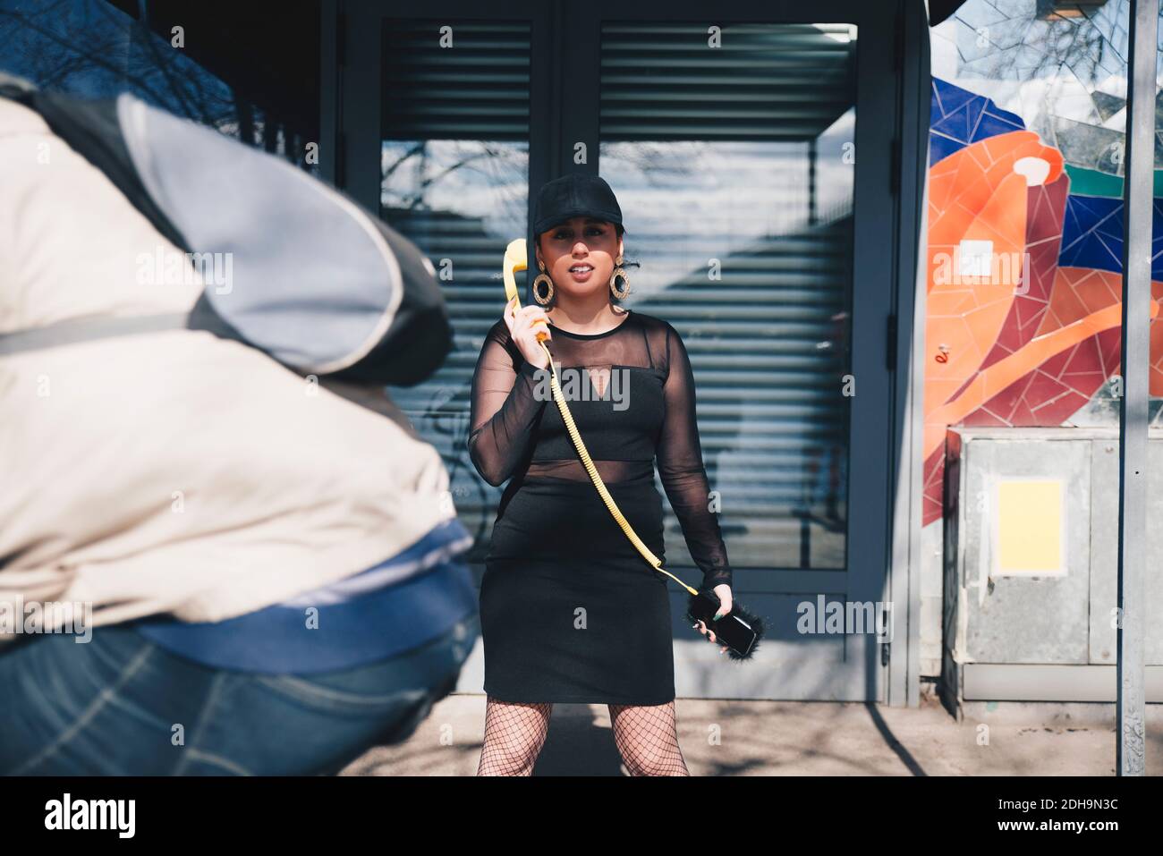 Porträt der jungen Frau hält Telefonhörer mit Handy verbunden Telefon, während Sie gegen das Gebäude stehen Stockfoto