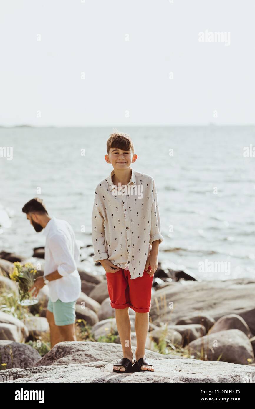Porträt eines lächelnden Jungen auf Felsen gegen Meer, während Vater Im Hintergrund stehend Stockfoto