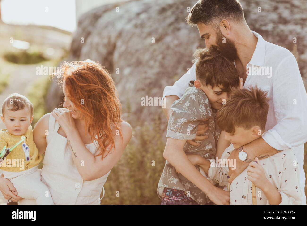 Lächelnder Vater umarmt Söhne, während Mutter auf Baby Mädchen Gegen Gesteinsbildung Stockfoto
