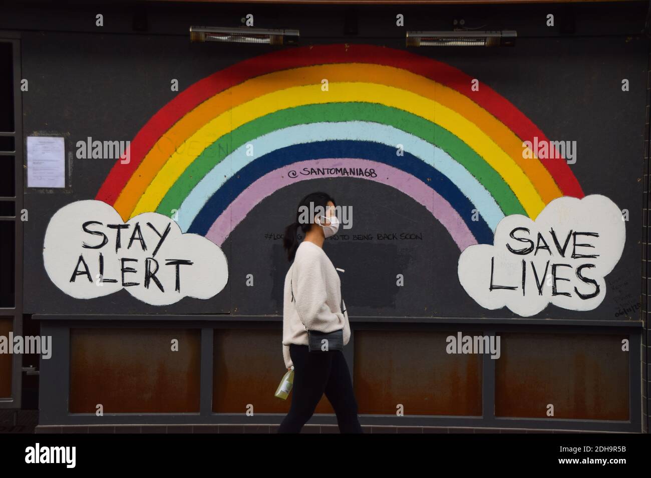 Eine Frau mit einer schützenden Gesichtsmaske geht an einem Stay Alert, Save Lives Regenbogenschild in einem geschlossenen Restaurant in der Old Compton Street in Soho, London vorbei. Stockfoto