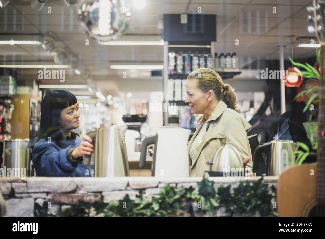 Verkäuferin mit Wasserkocher beim Betrachen der Kundin im Geschäft gesehen Durch Glasfenster Stockfoto