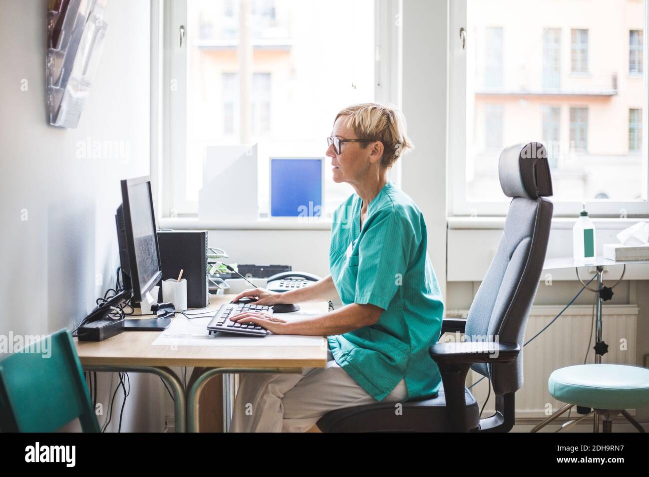 Seitenansicht der reifen weiblichen Arzt arbeiten über Computer, während Sitzen in der Klinik Stockfoto