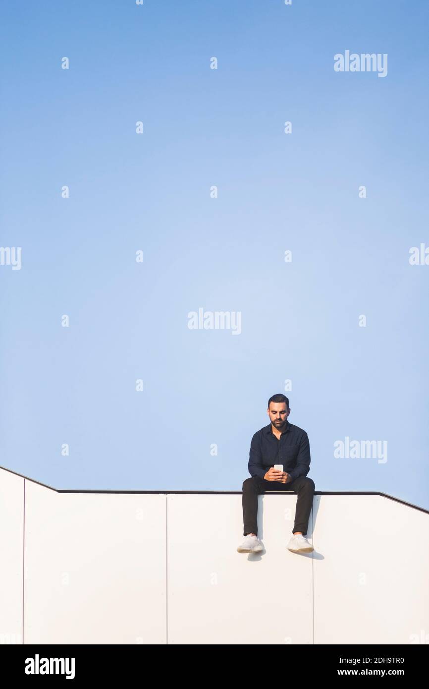 Mann mit Telefon, während auf gebaute Struktur gegen blau sitzen Himmel Stockfoto