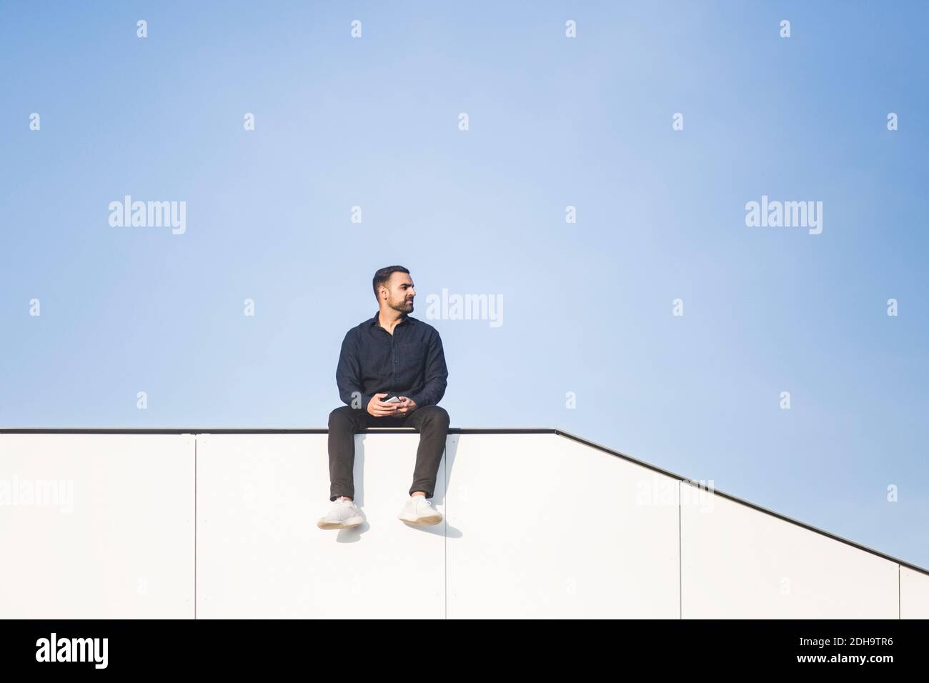 Betrachtend Mann wegschauen, während er auf gebauter Struktur gegen sitzt Blauer Himmel Stockfoto