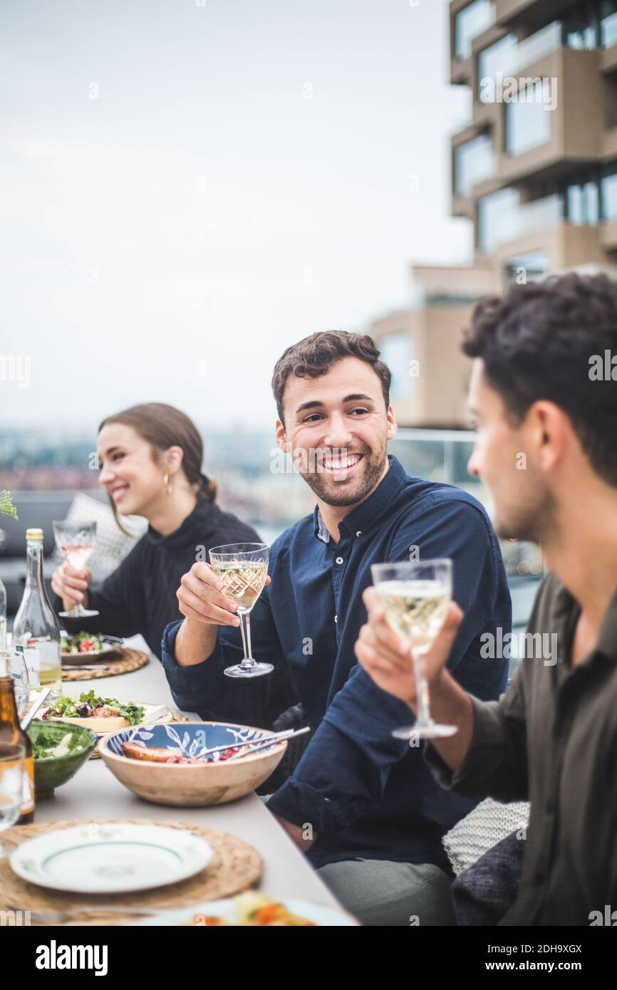 Lächelnder Mann, der mit einem Freund spricht, während er von einer Frau sitzt Party auf dem Dach Stockfoto