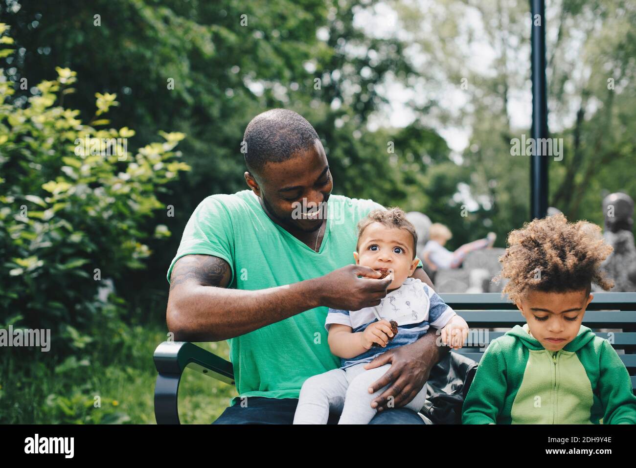Lächelnder Vater Fütterung Baby Junge während sitzen mit Sohn auf Bank im Park Stockfoto Lächelnder Vater Fütterung Baby Junge während sitzen mit Sohn auf Bank im Park Stockfoto