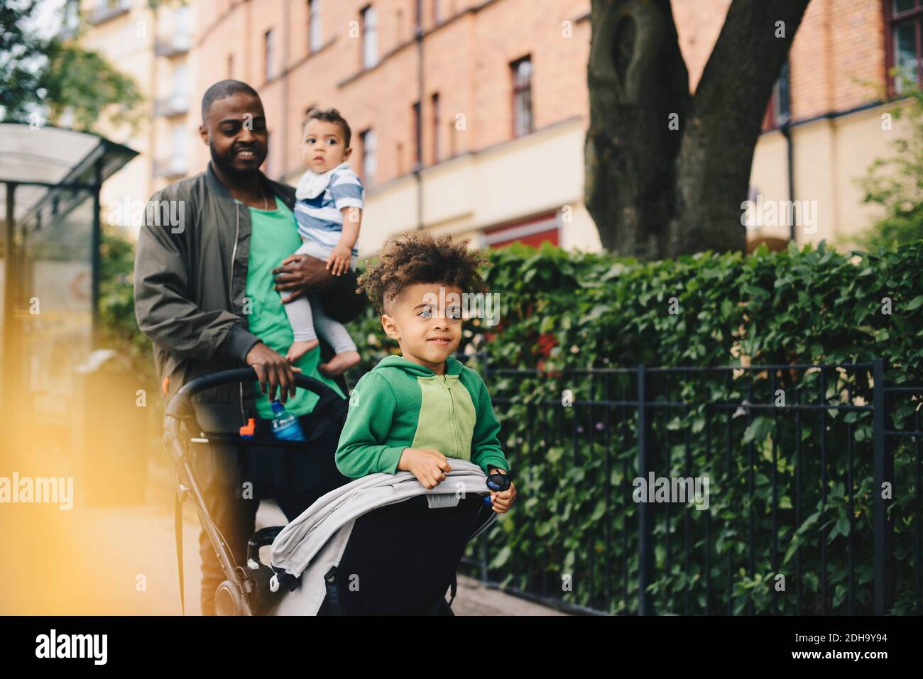 Vater schob Kinderwagen, während er mit Kindern am Zaun spazieren ging In der Stadt Stockfoto