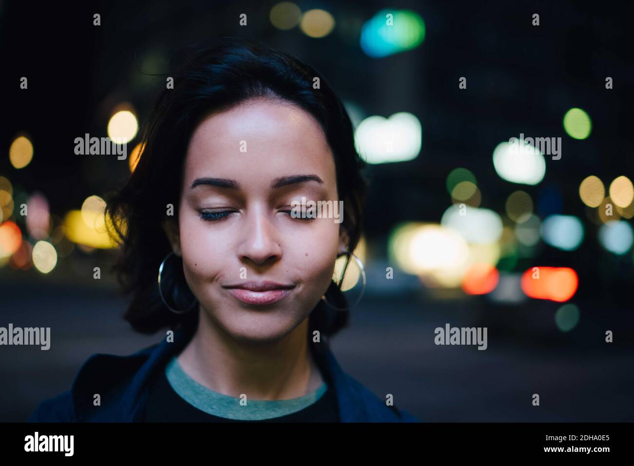 Lächelnde Frau mit geschlossenen Augen stehend in der beleuchteten Stadt an Nacht Stockfoto
